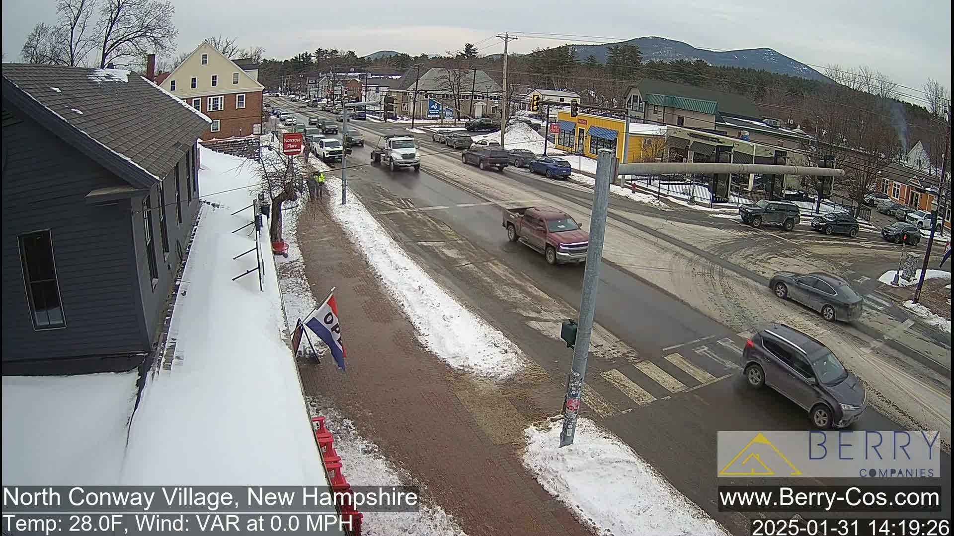 A snow-covered street scene shows several vehicles driving through a town on a partly cloudy day.
