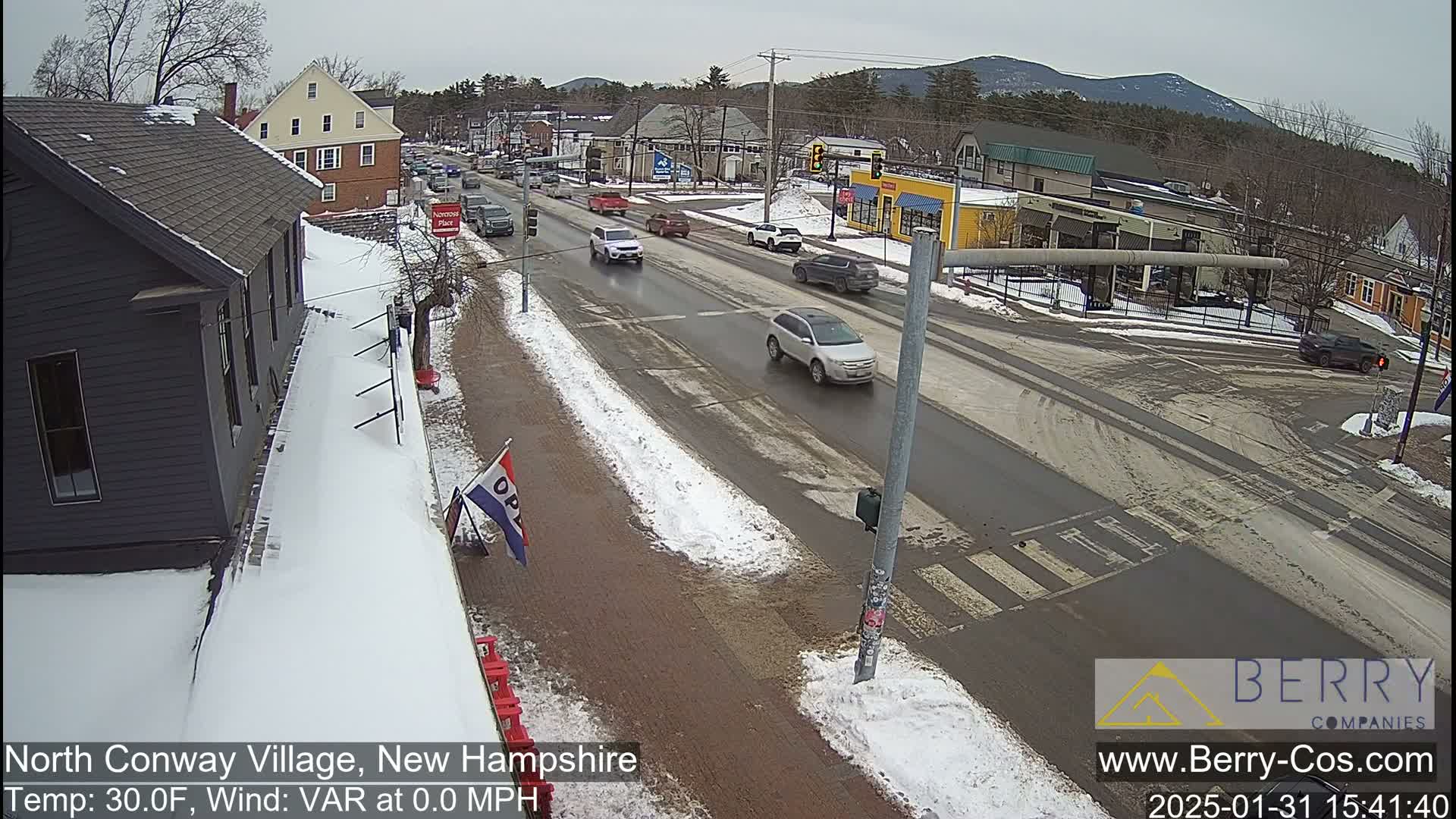 A partly snowy street scene shows several cars driving on a road that intersects with another road, with buildings and mountains visible in the background under a cloudy sky.