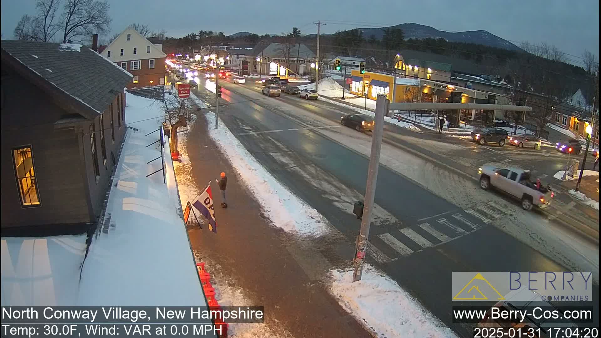 A snow-covered street scene in a village shows cars driving past buildings under a twilight sky.