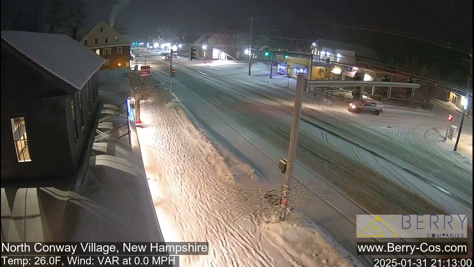 A snow-covered street scene at night shows a few cars driving past buildings under lightly falling snow.