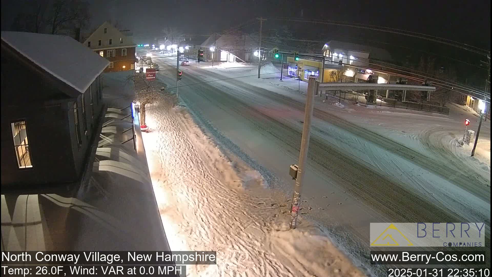 A snow-covered street scene at night shows a few buildings and cars under lightly falling snow.