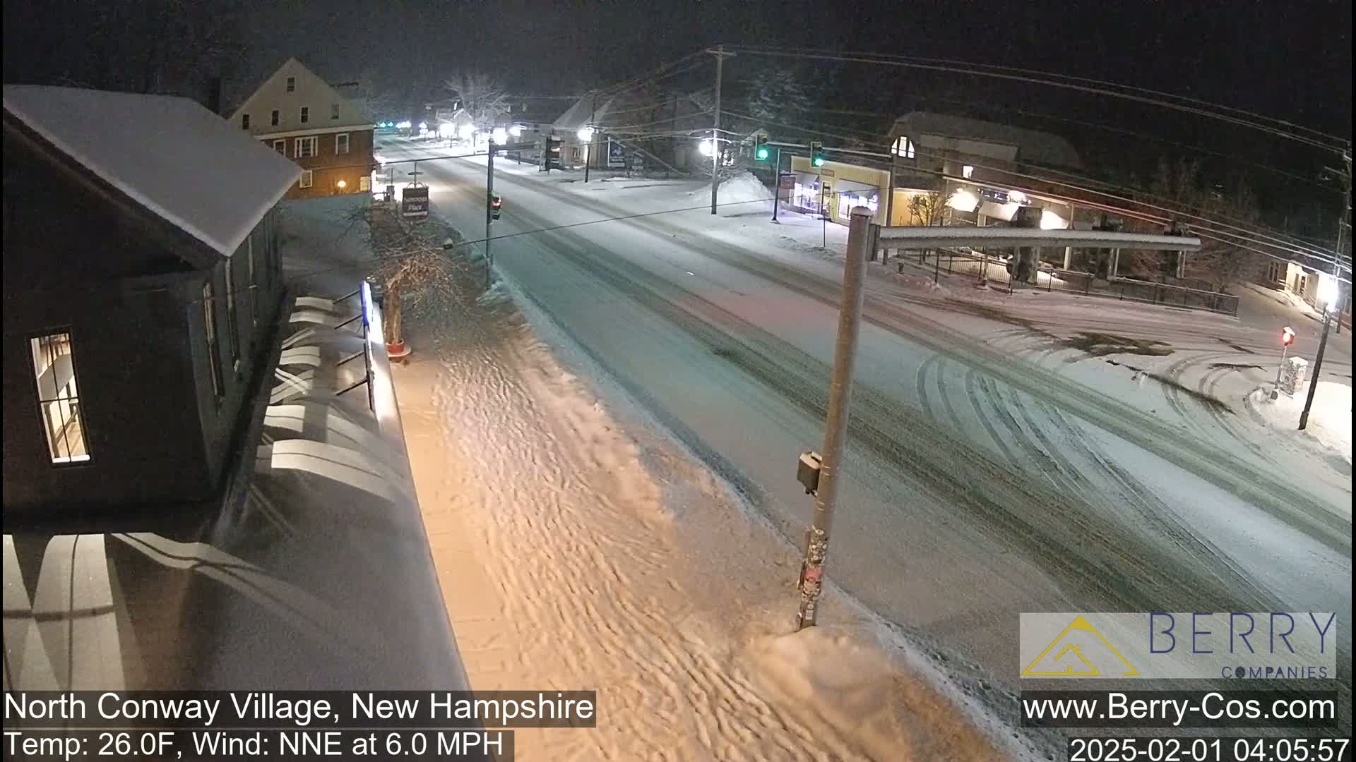 A snow-covered street scene at night, with buildings and vehicles visible under a light snowfall and a temperature of 26°F, with a northeast wind at 6.0 mph.