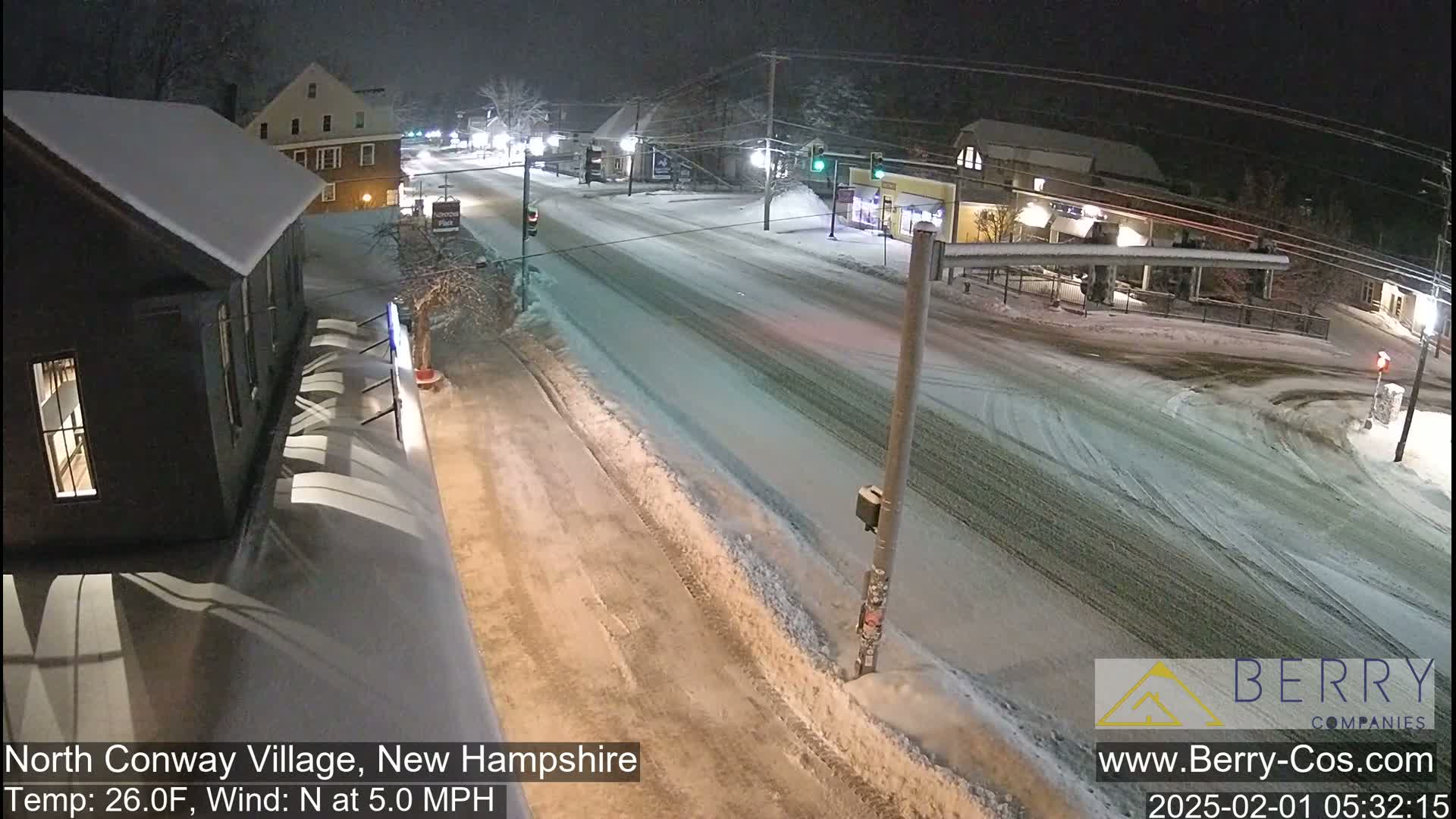 A snow-covered street scene at night shows a road intersection with buildings and sparse traffic under a lightly falling snow.