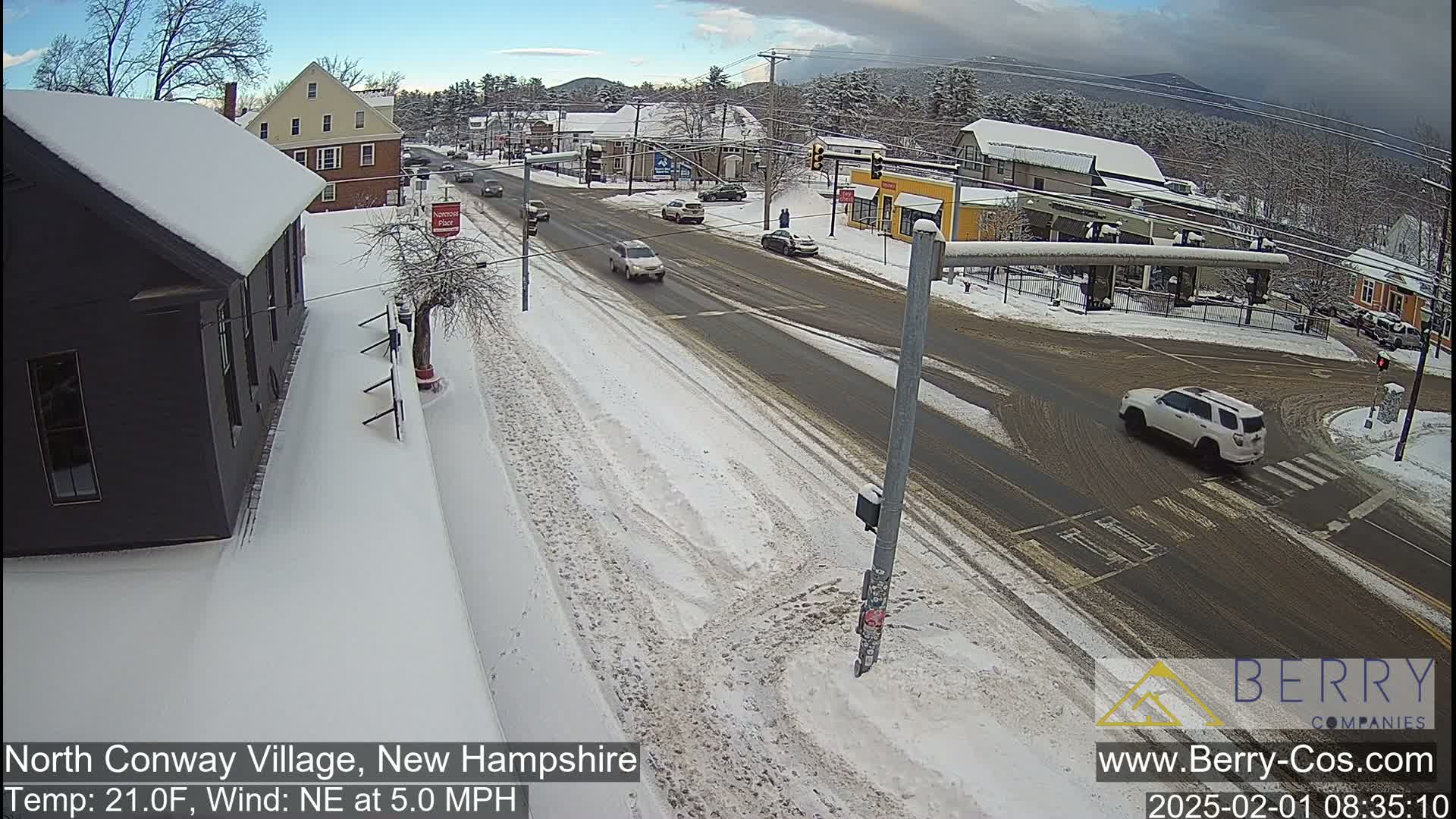 A snow-covered street scene shows several cars driving through a town intersection under a partly cloudy sky.