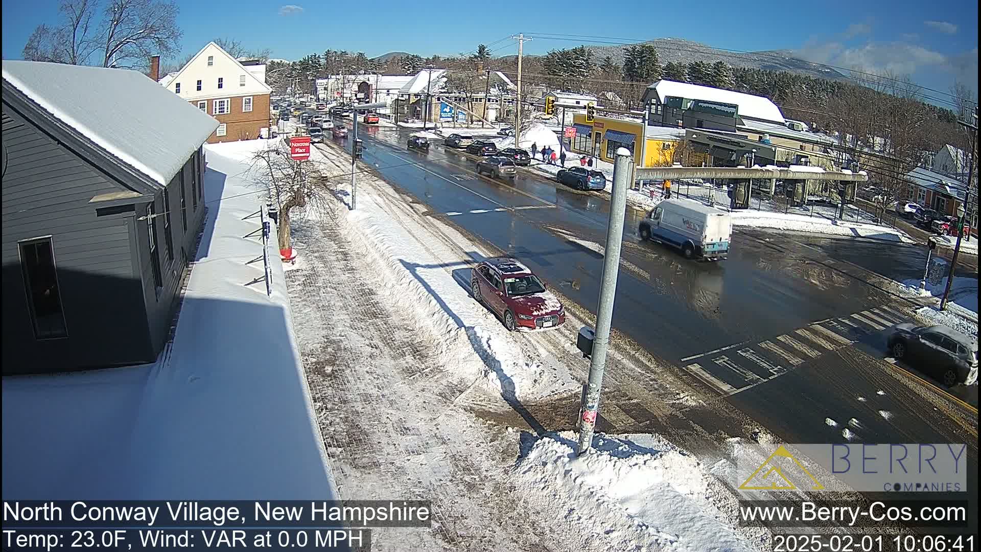 A snow-covered street scene shows several cars driving on a wet road with patches of snow and ice, under a sunny sky.