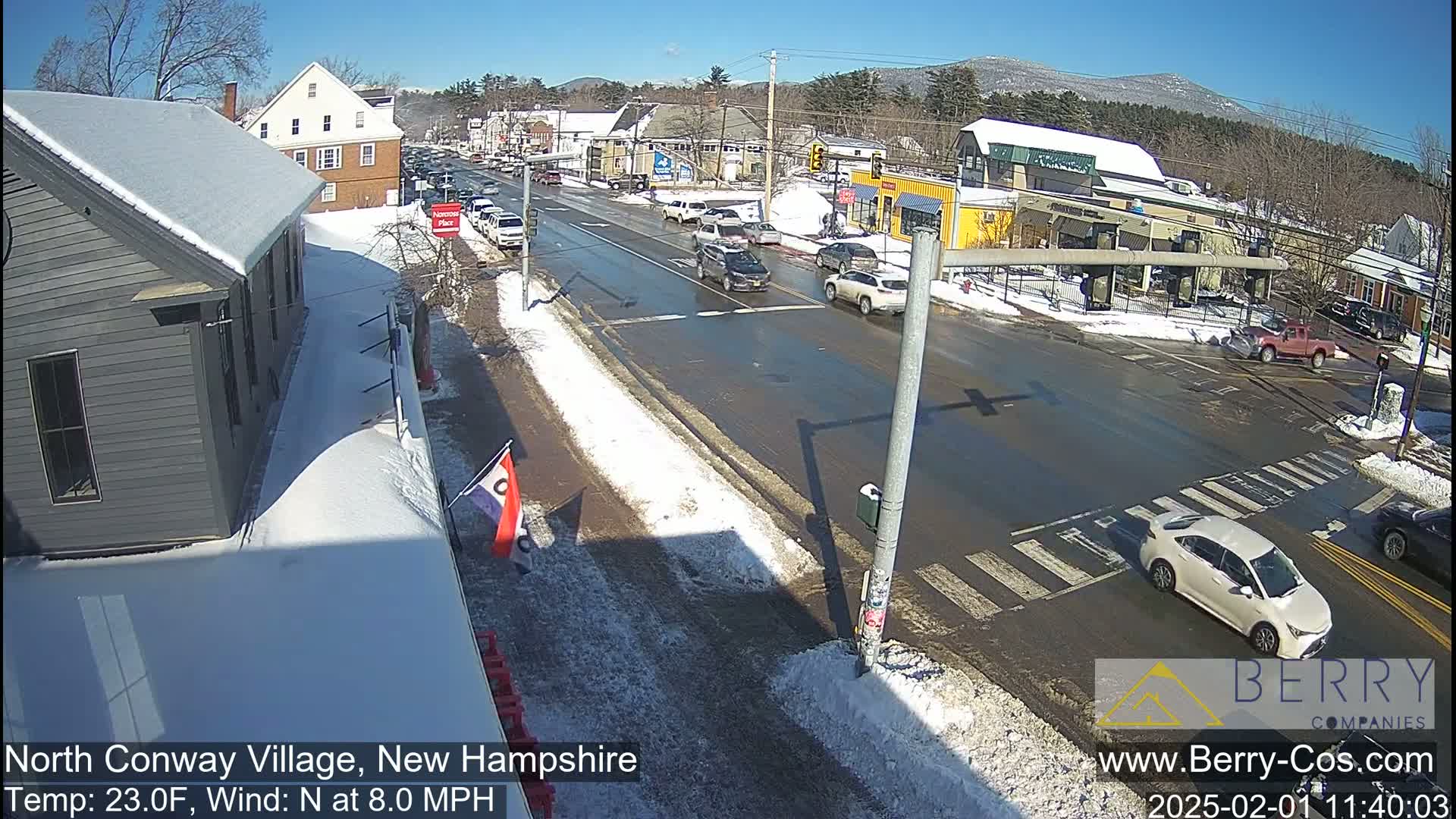 A snow-covered street scene in a small town on a sunny day with a north wind, featuring several cars at an intersection and buildings on either side of the road.