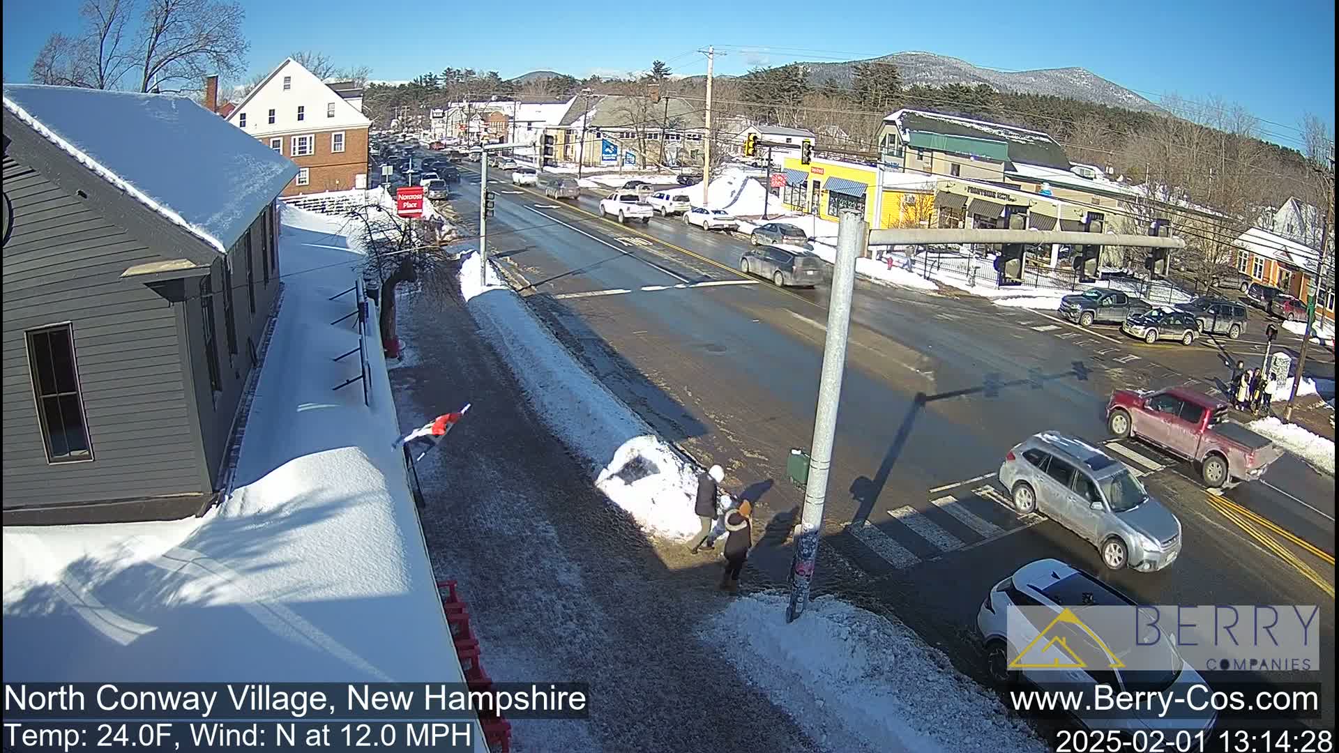 A snow-covered street scene on a sunny day shows several vehicles at an intersection, with pedestrians walking near a snow bank in front of buildings.