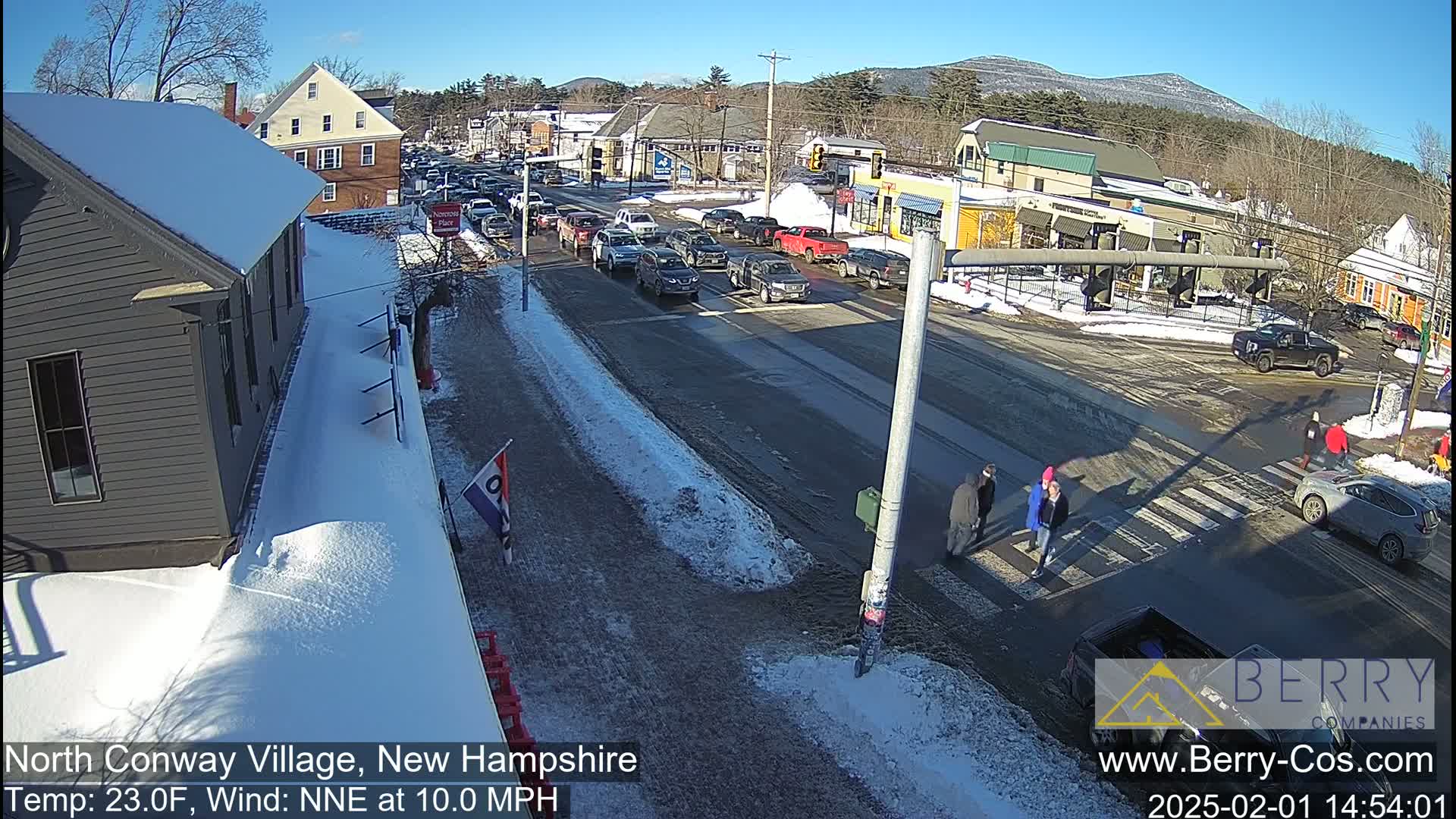 A sunny, snow-covered town intersection shows several cars stopped at a crosswalk where pedestrians are walking, with a partially snow-covered street and buildings in the background.