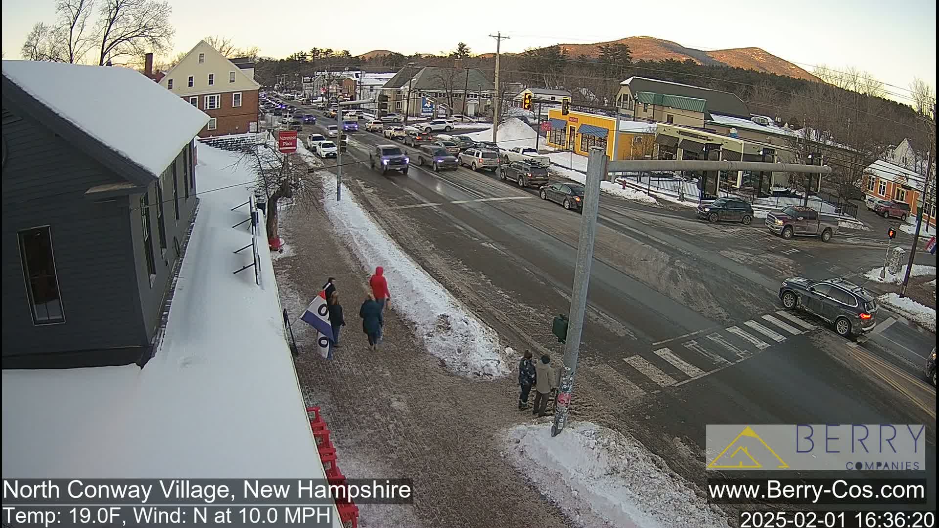 A snowy street scene shows a partly cloudy sky, moderate traffic, and pedestrians walking near a crossroads with several buildings in the background.