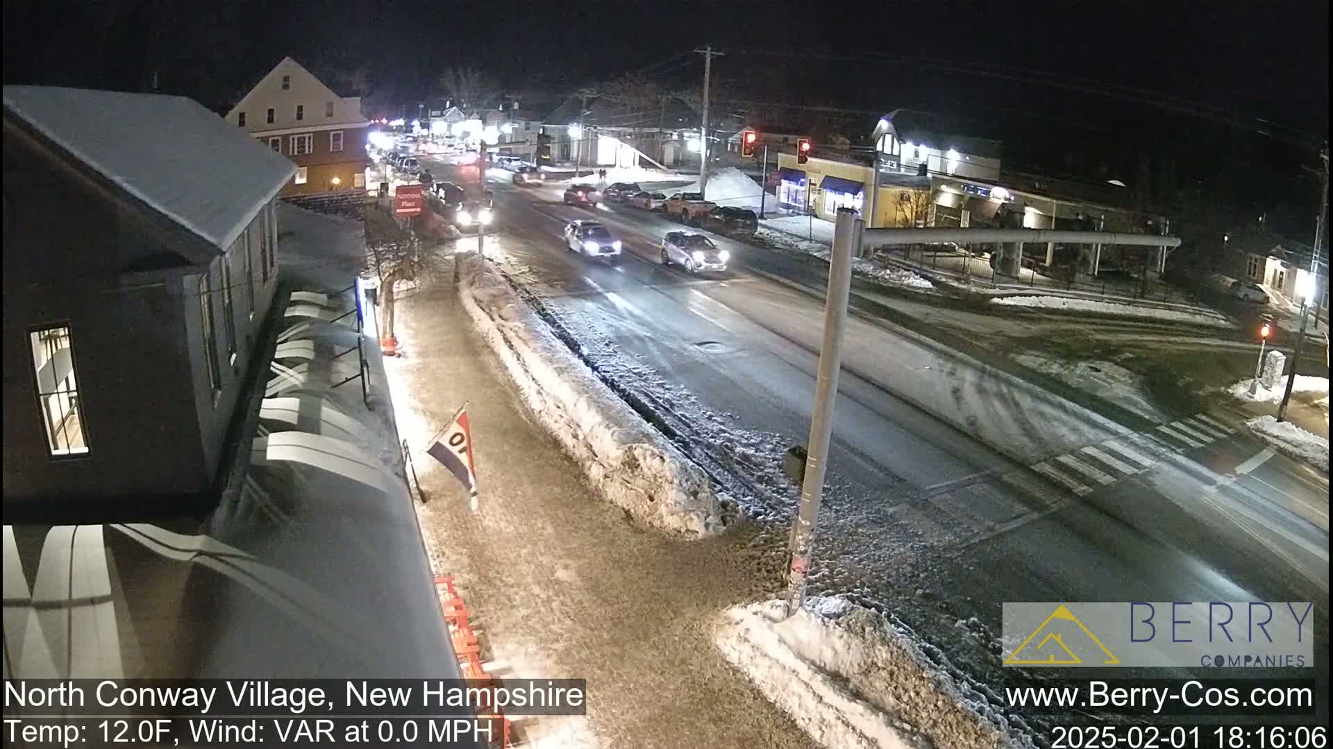 A snow-covered street scene at night shows several cars driving on a road, past buildings with snow on their roofs.