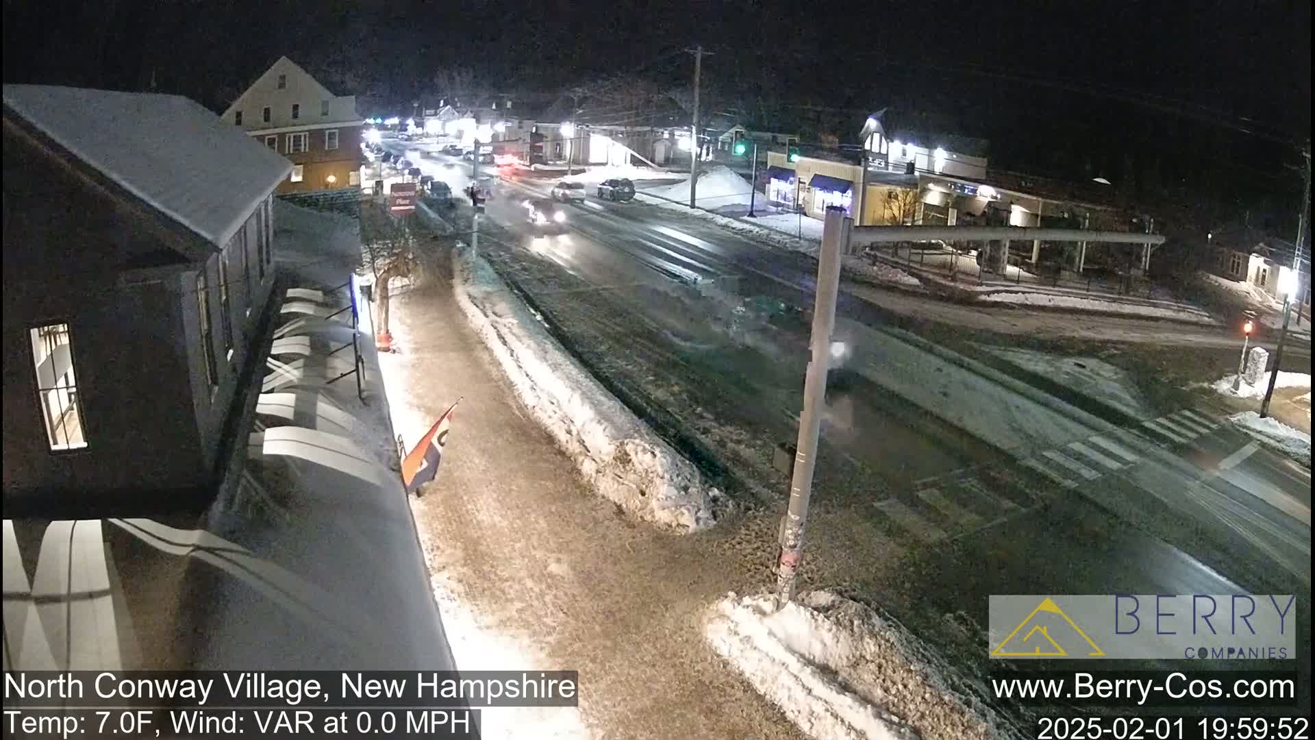 A nighttime, snow-covered street scene shows several vehicles traveling on a road, with buildings and snow-covered sidewalks on either side under a dark sky.