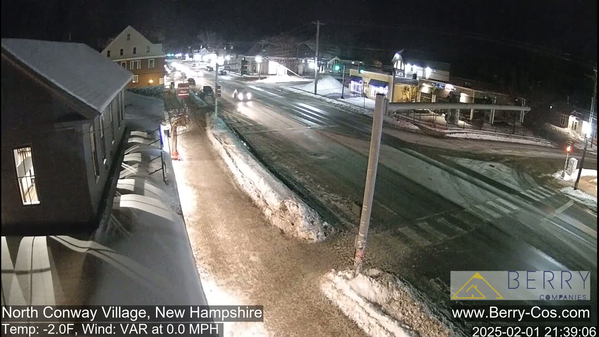 A snow-covered street intersection at night with a few cars visible, under dark, clear skies.