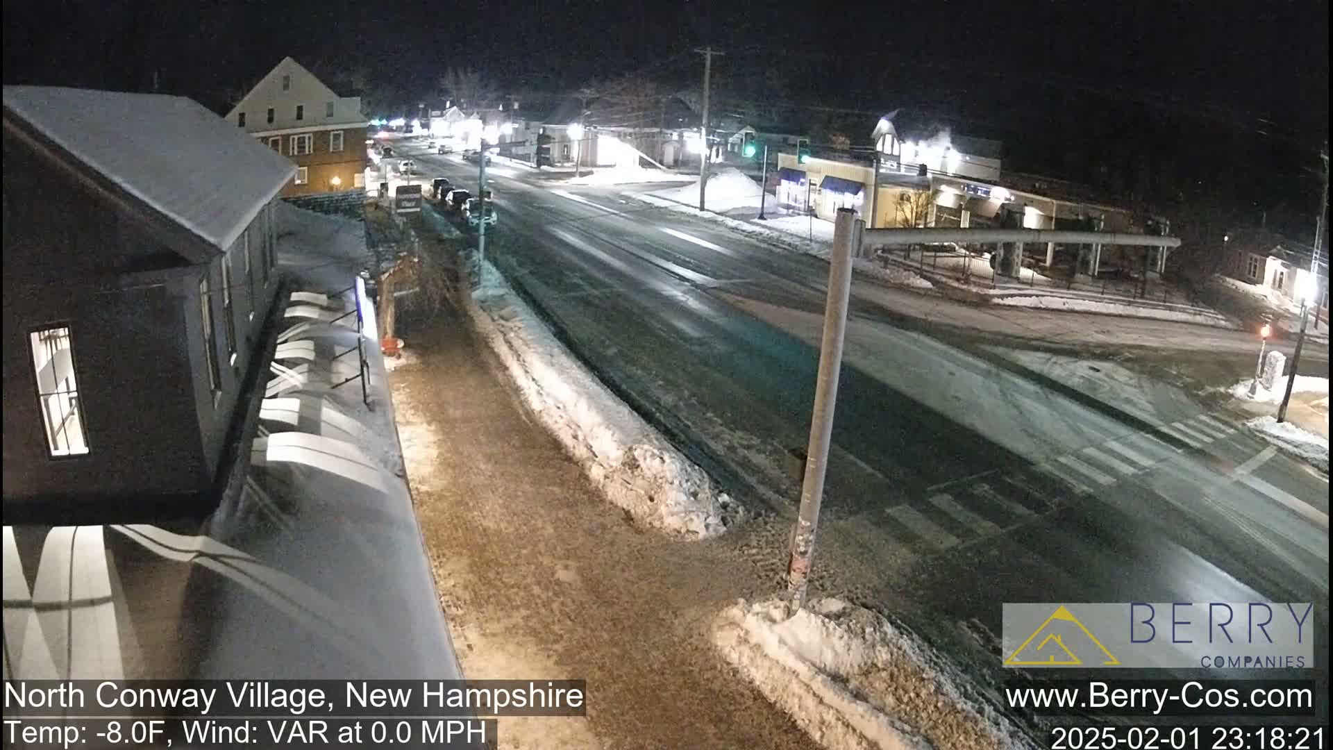 A snow-covered street scene at night shows a quiet intersection in a small town with buildings lining the road.