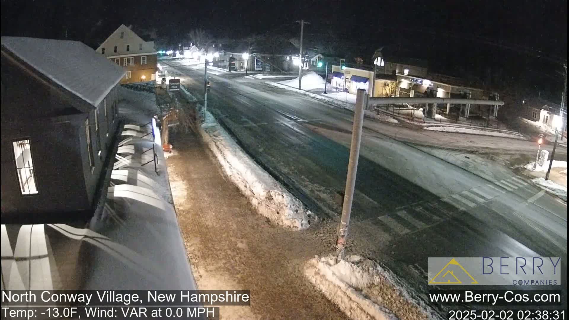 A snow-covered street scene at night shows a mostly empty road with buildings and some parked cars under a dark sky.