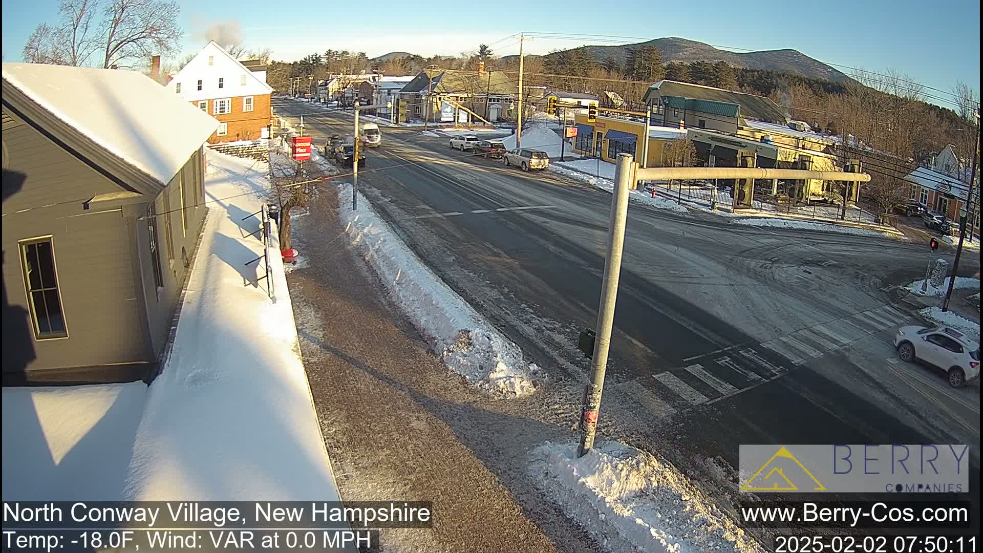 A sunny, snowy street scene shows a road intersection in a small town, with cars driving and snow piled on the sides of the road.