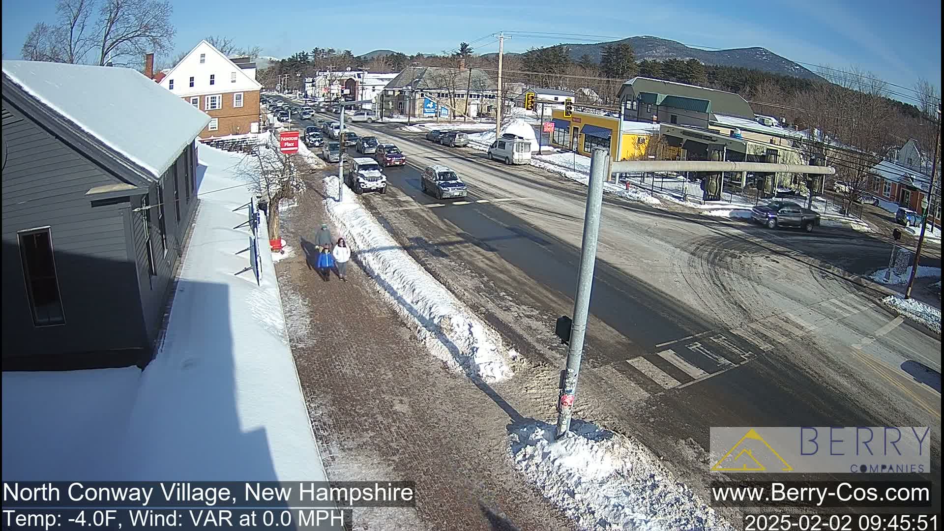 A sunny winter day shows a town intersection with cars and pedestrians, snow-covered sidewalks, and mountains in the background.