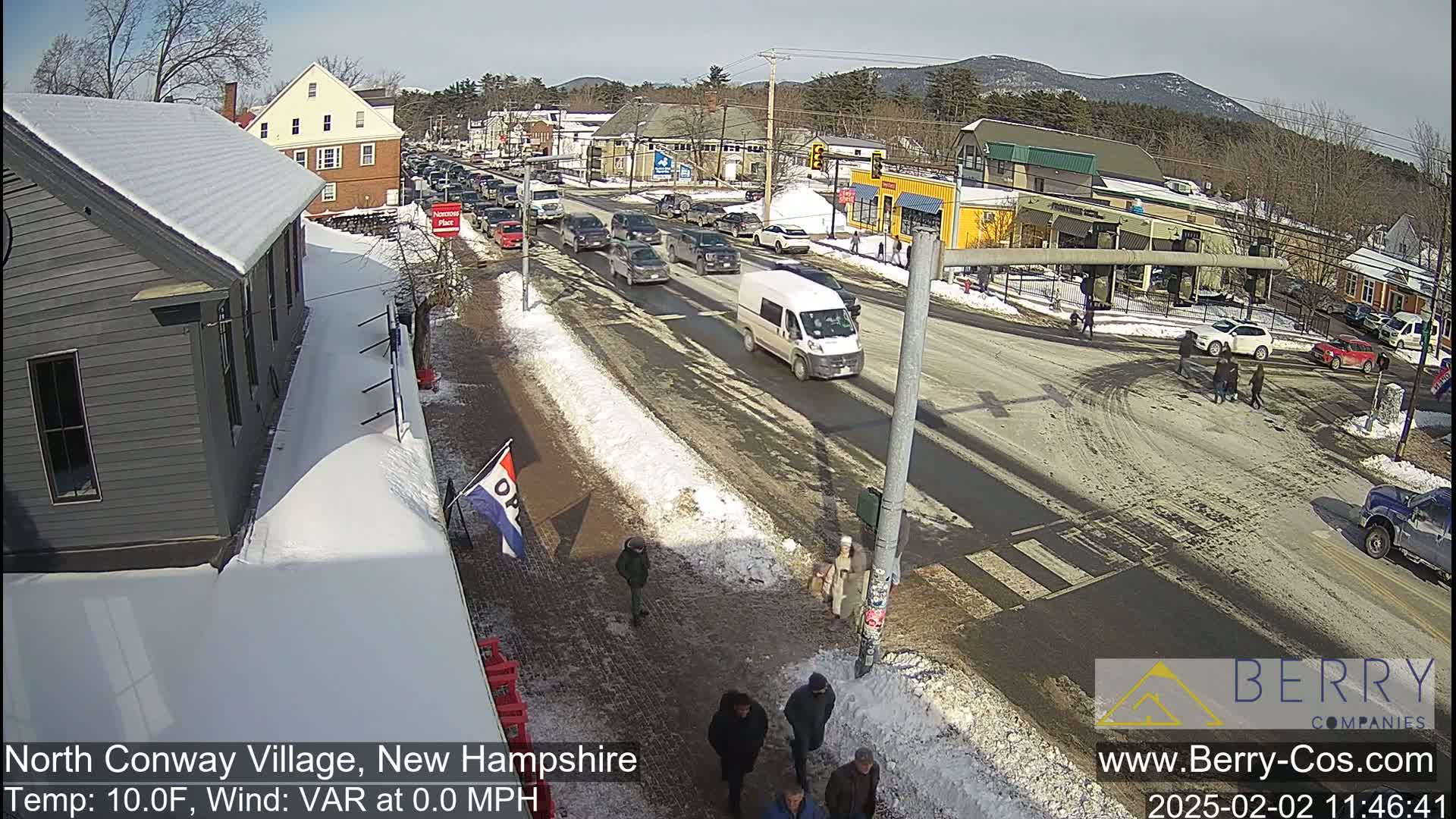 A sunny winter day shows a partly snow-covered street intersection in a village, with cars driving and pedestrians walking, under a clear sky.