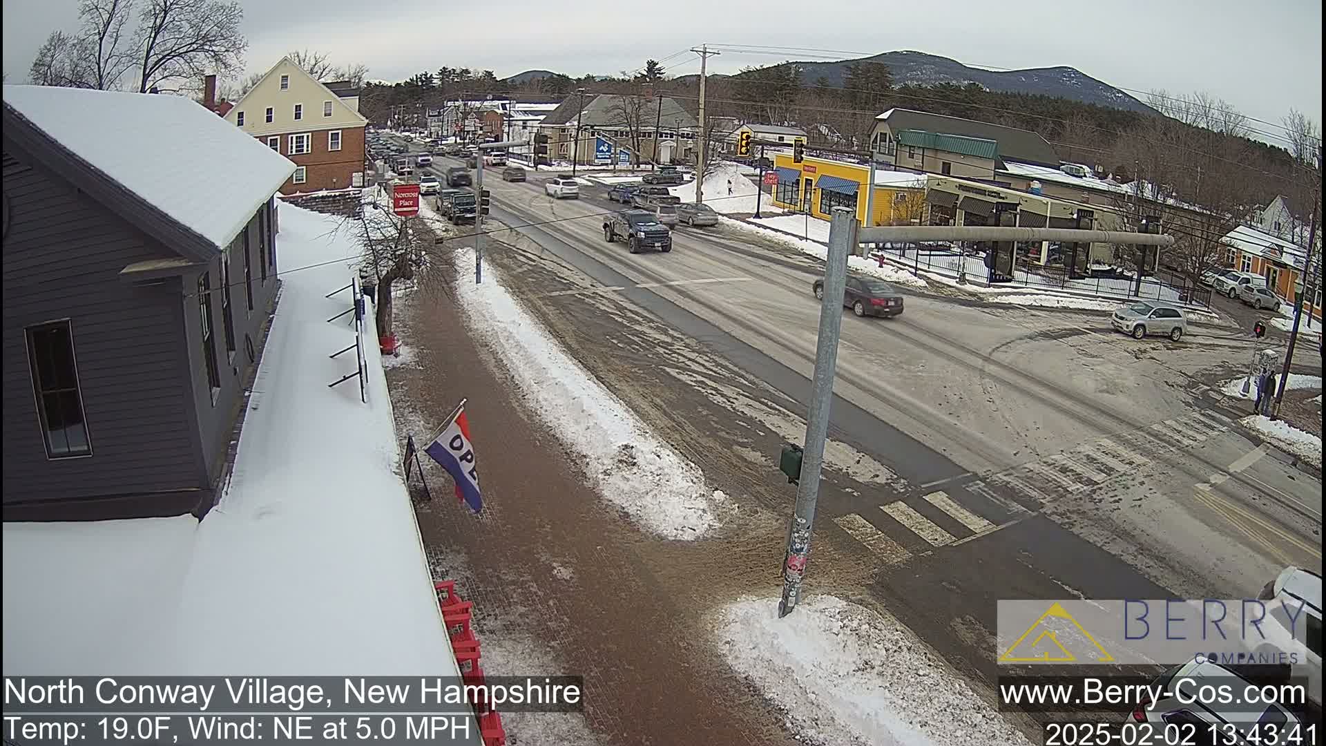 A snow-covered street scene in a small town shows several cars driving on a main road, with some buildings and patches of snow along the sidewalks, under an overcast sky.
