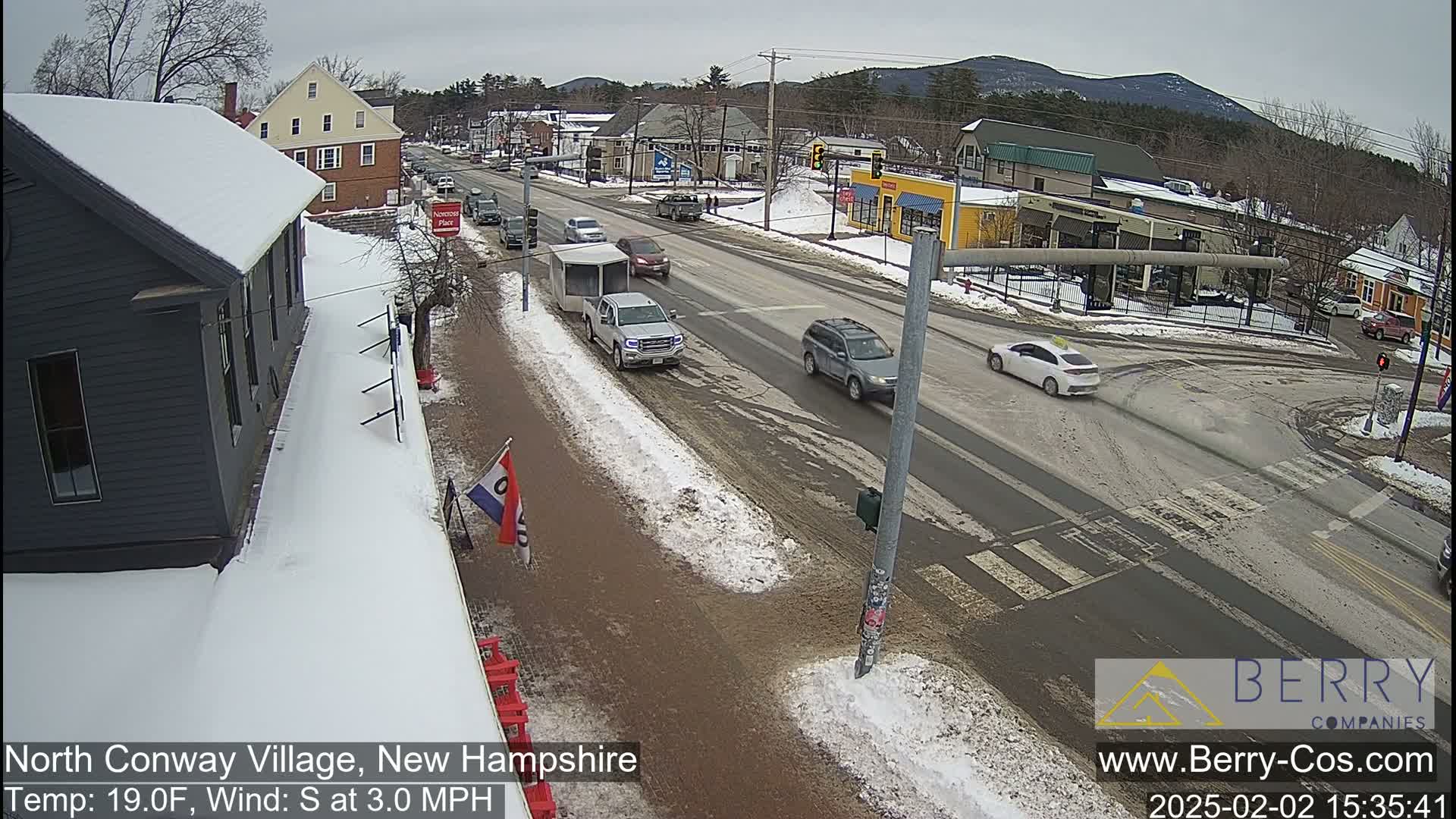 A street intersection in a small town with snow on the ground and some vehicles driving, under a cloudy sky.