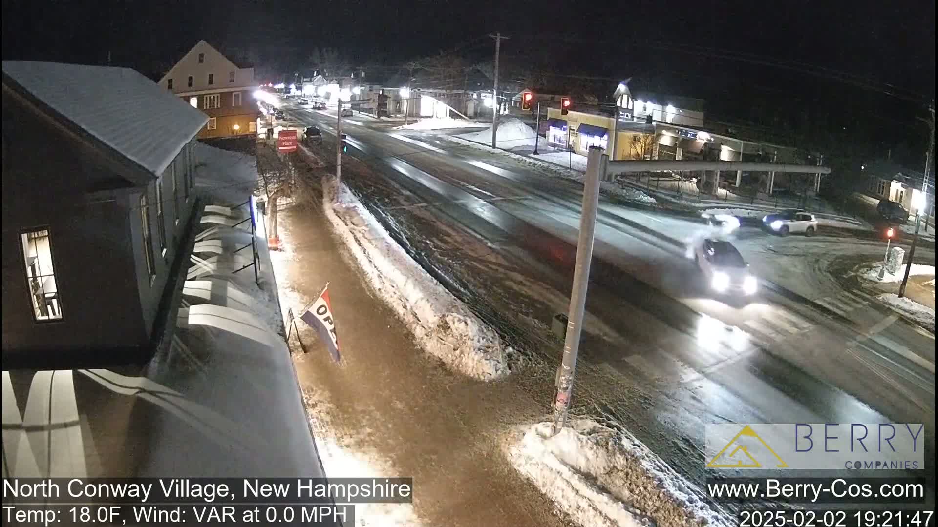 A nighttime, snow-covered street intersection with several vehicles, buildings, and streetlights visible under calm, cold conditions.