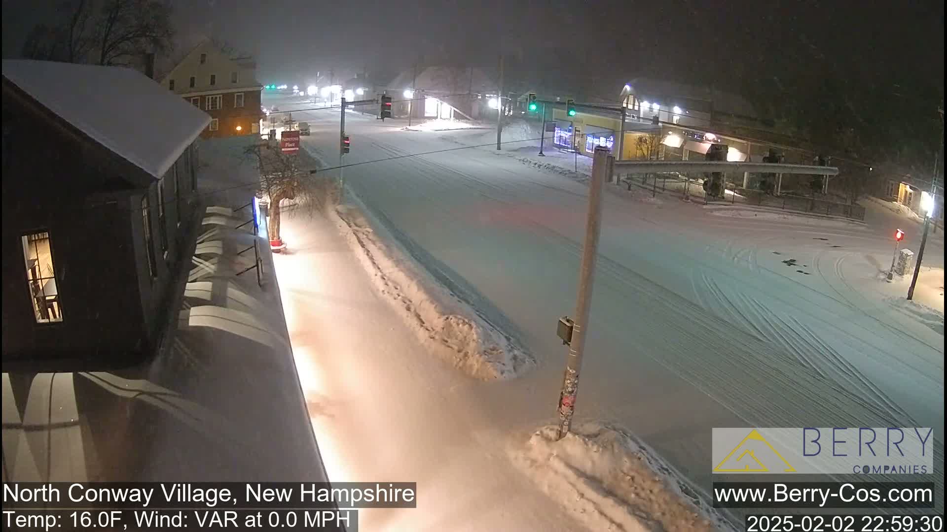 A snow-covered street scene at night shows a crossroads with buildings and streetlights under light snowfall with calm winds.