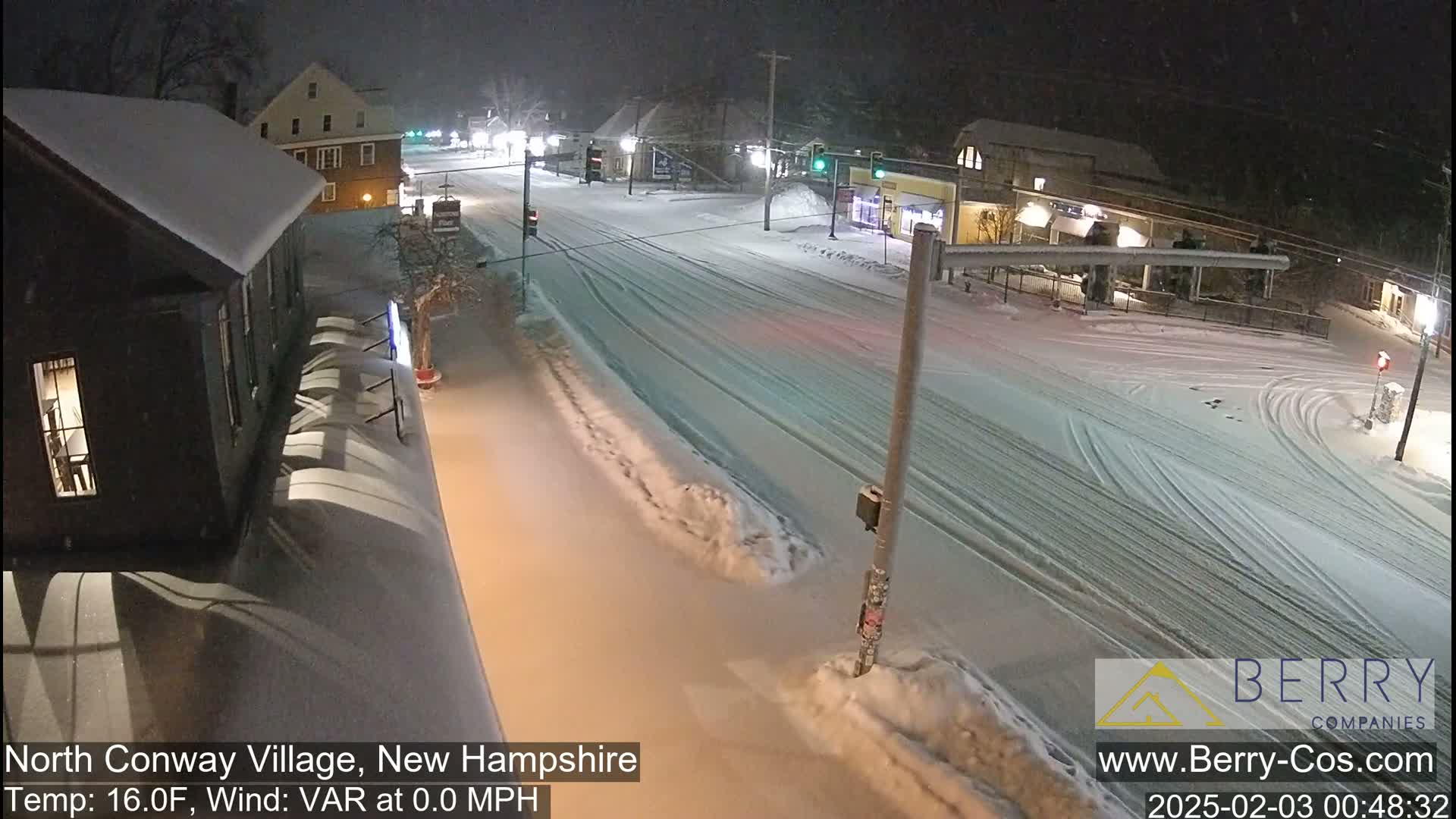 A snow-covered street scene at night shows several buildings and vehicles with tire tracks in the snow.