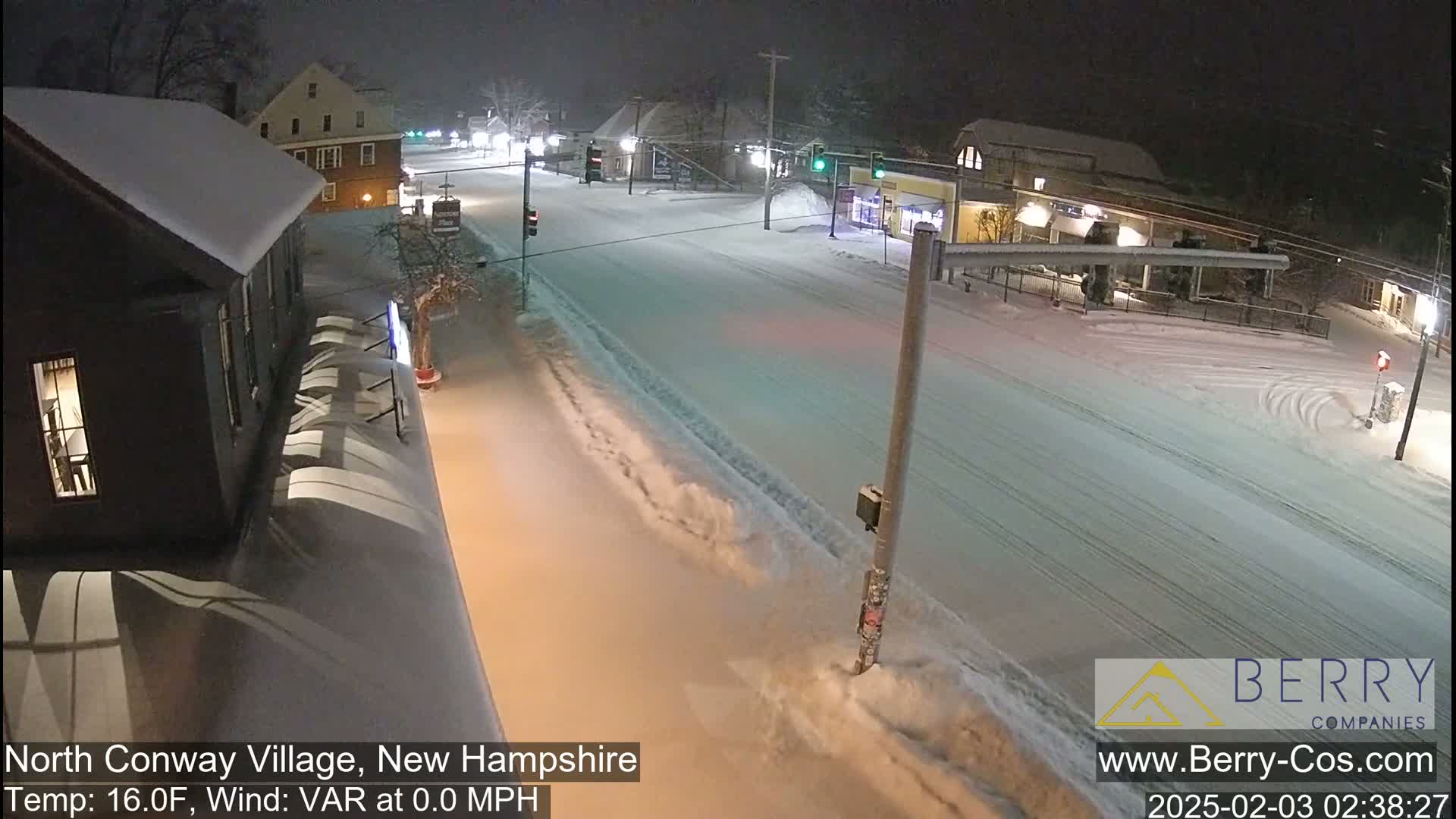 A snow-covered street scene at night shows buildings with snow-laden roofs, under calm, snowy conditions.