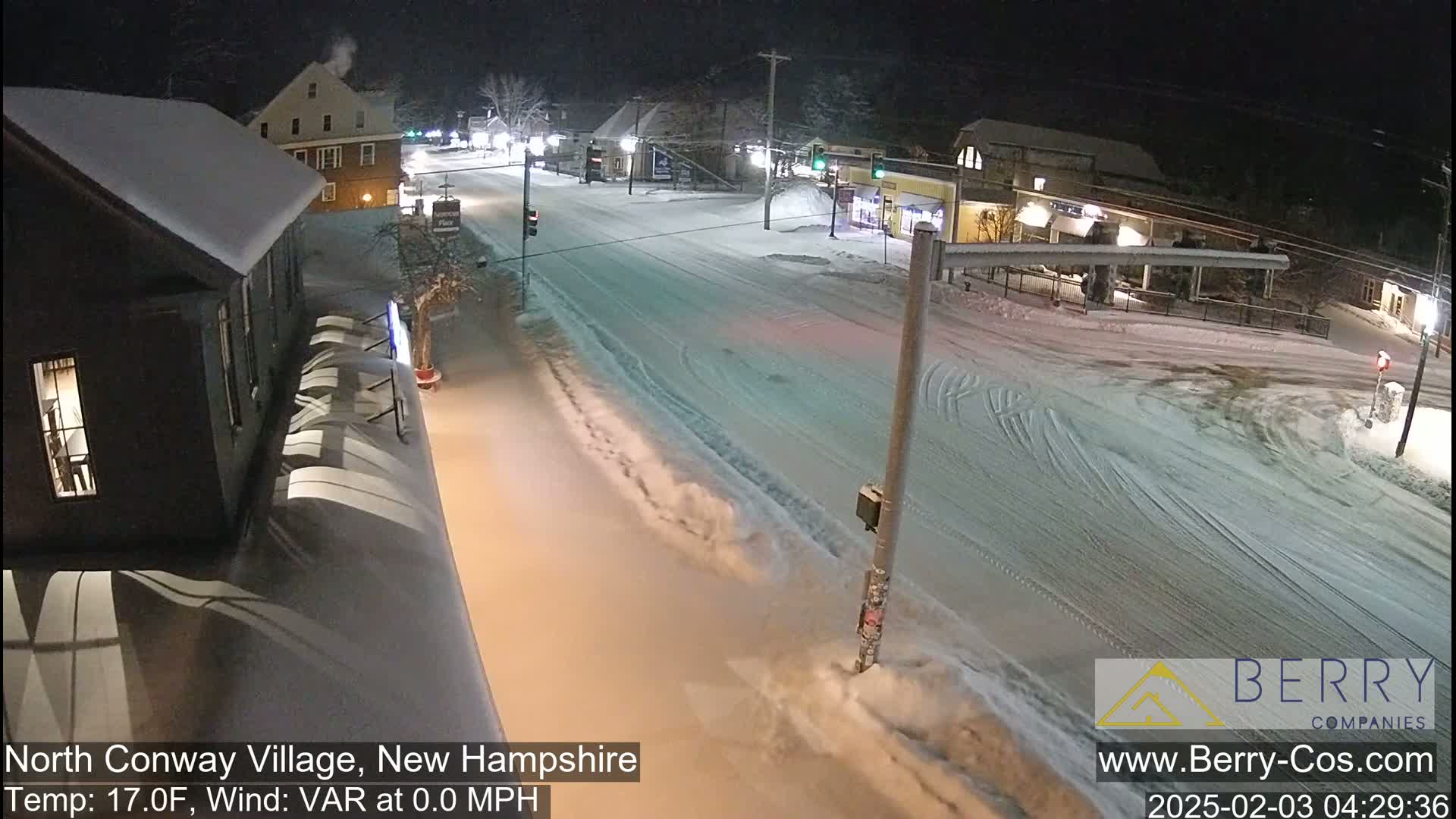 A snow-covered street scene at night shows buildings with snow on their roofs and tire tracks in the snow on the road.
