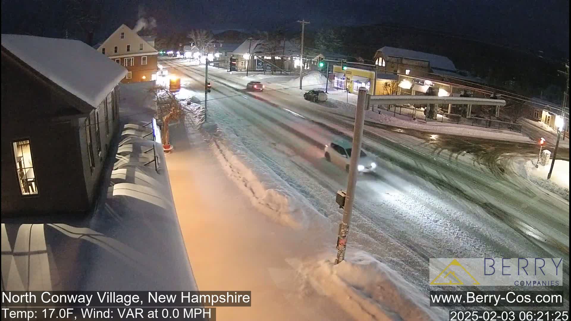 A snow-covered street scene at night shows several cars driving on a two-lane road past buildings with snow-covered roofs.