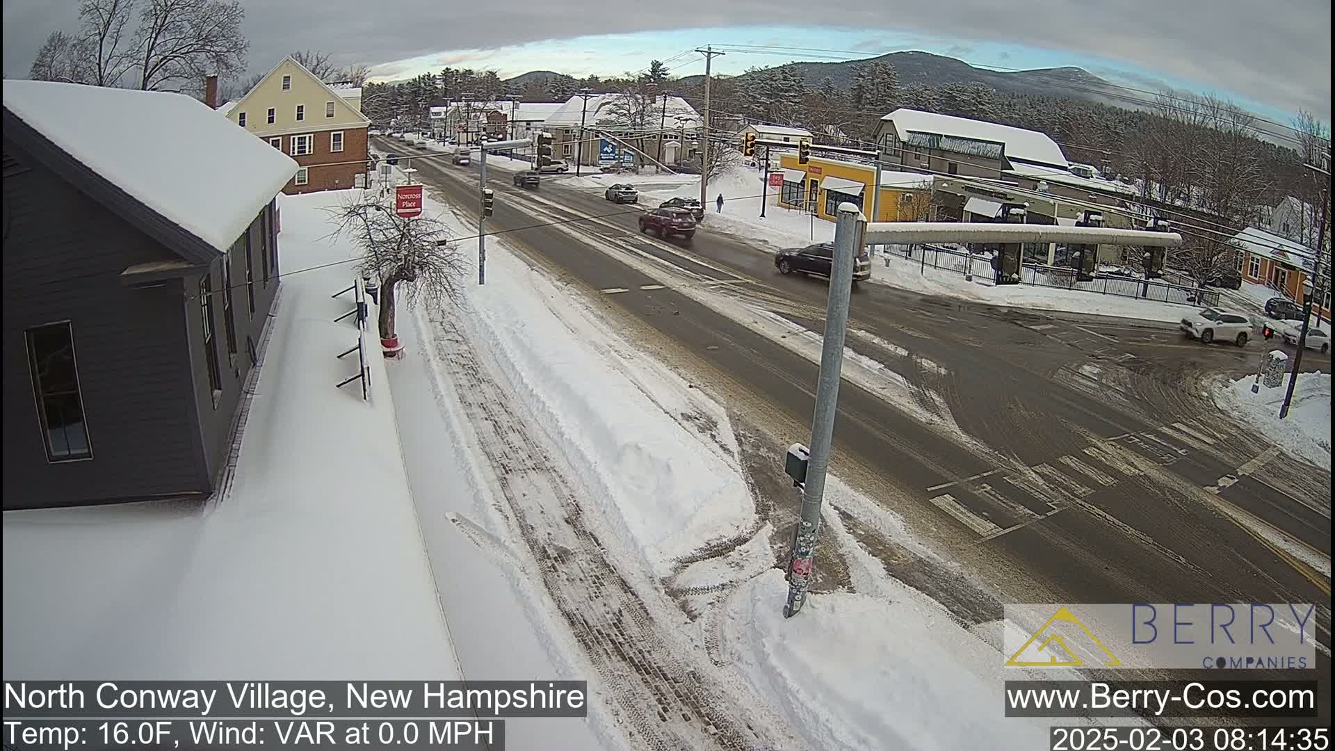 A snow-covered street scene shows a four-way intersection with several cars and buildings, under a partly cloudy sky.