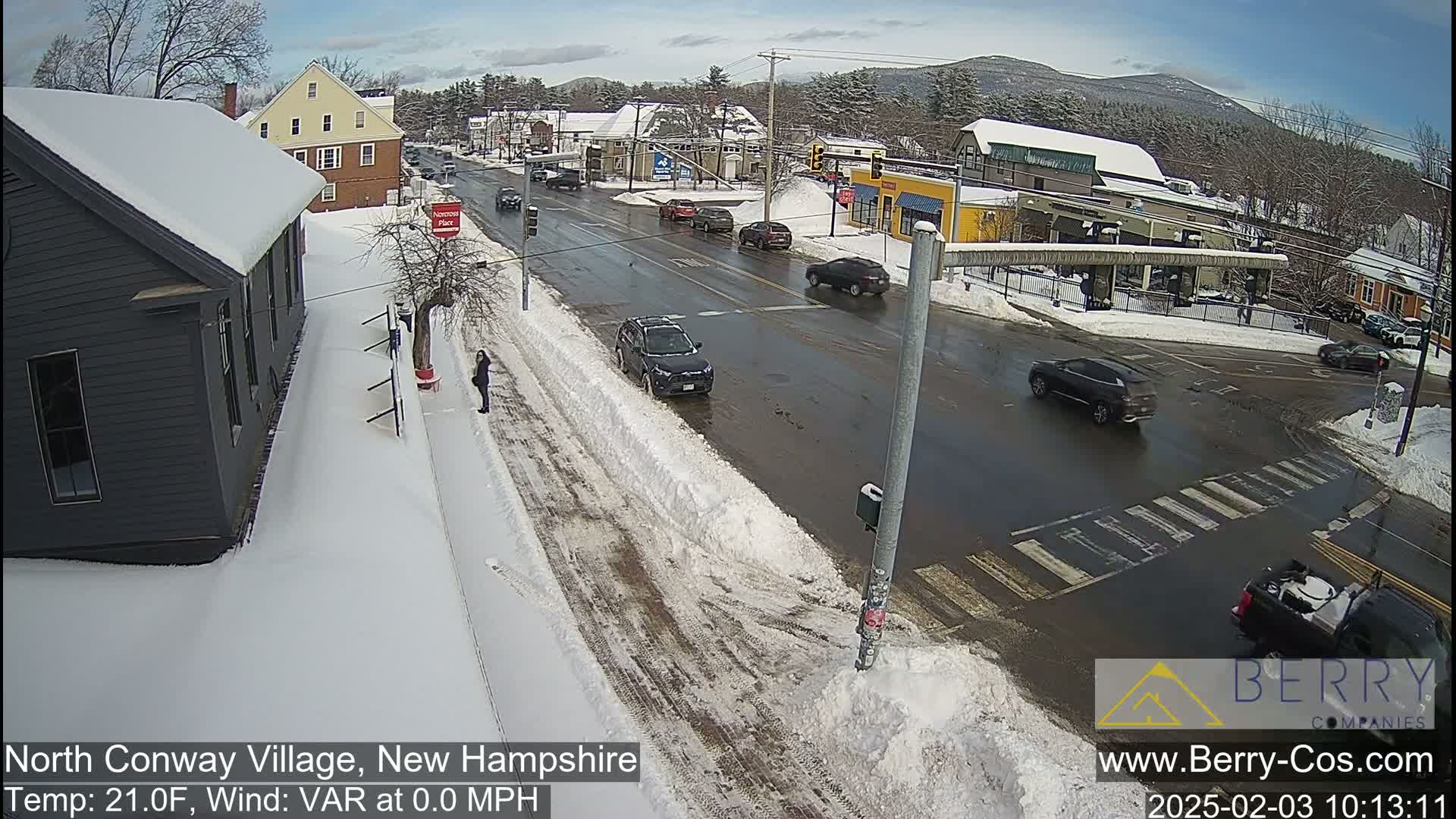 A snow-covered street intersection in a small town, with several cars and pedestrians present under a clear, sunny sky.