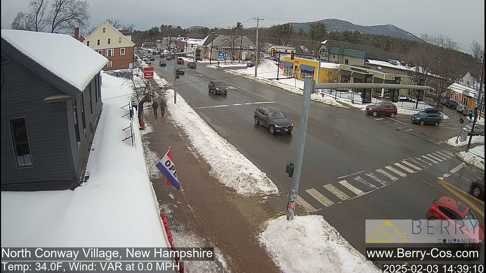 A snow-covered street scene shows several cars at an intersection in a village, with a partly cloudy sky above.