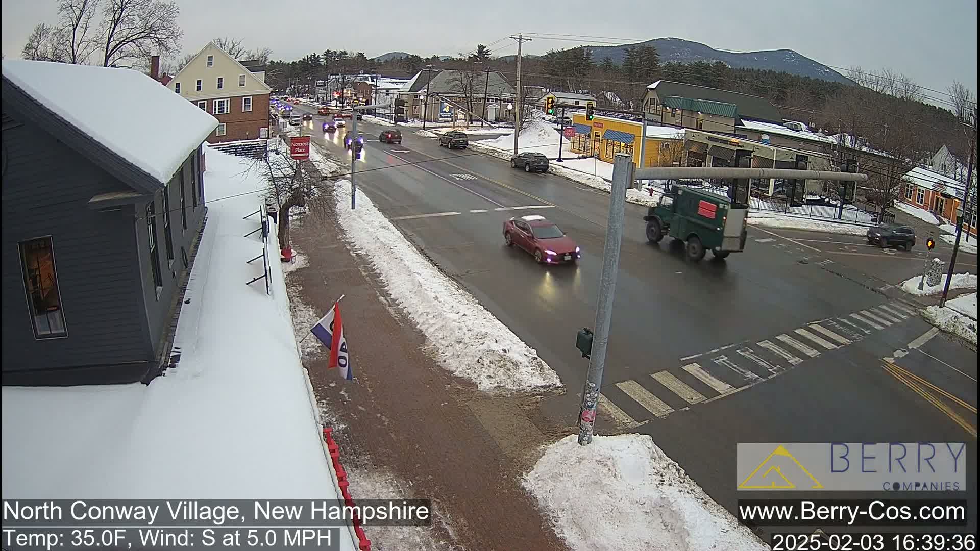 A partly snowy street scene shows several vehicles at an intersection in a village setting under a cloudy sky.