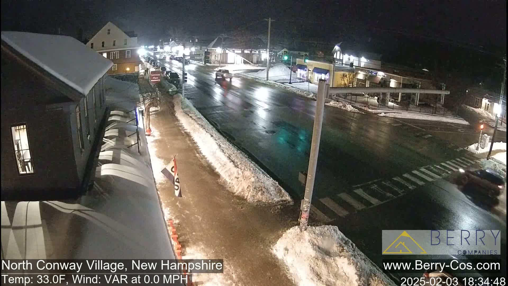 A nighttime street scene shows a snow-covered road with a few cars driving on it, under calm conditions.