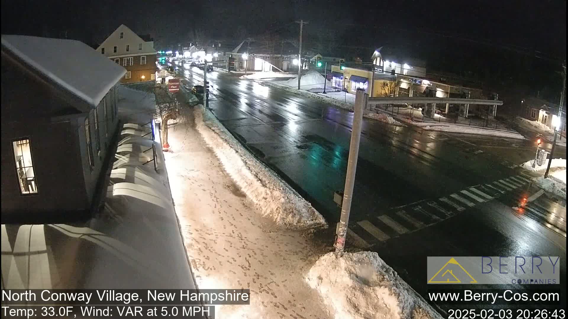 A nighttime street scene shows a wet road with snow banks and footprints on the sidewalk under a dark sky.