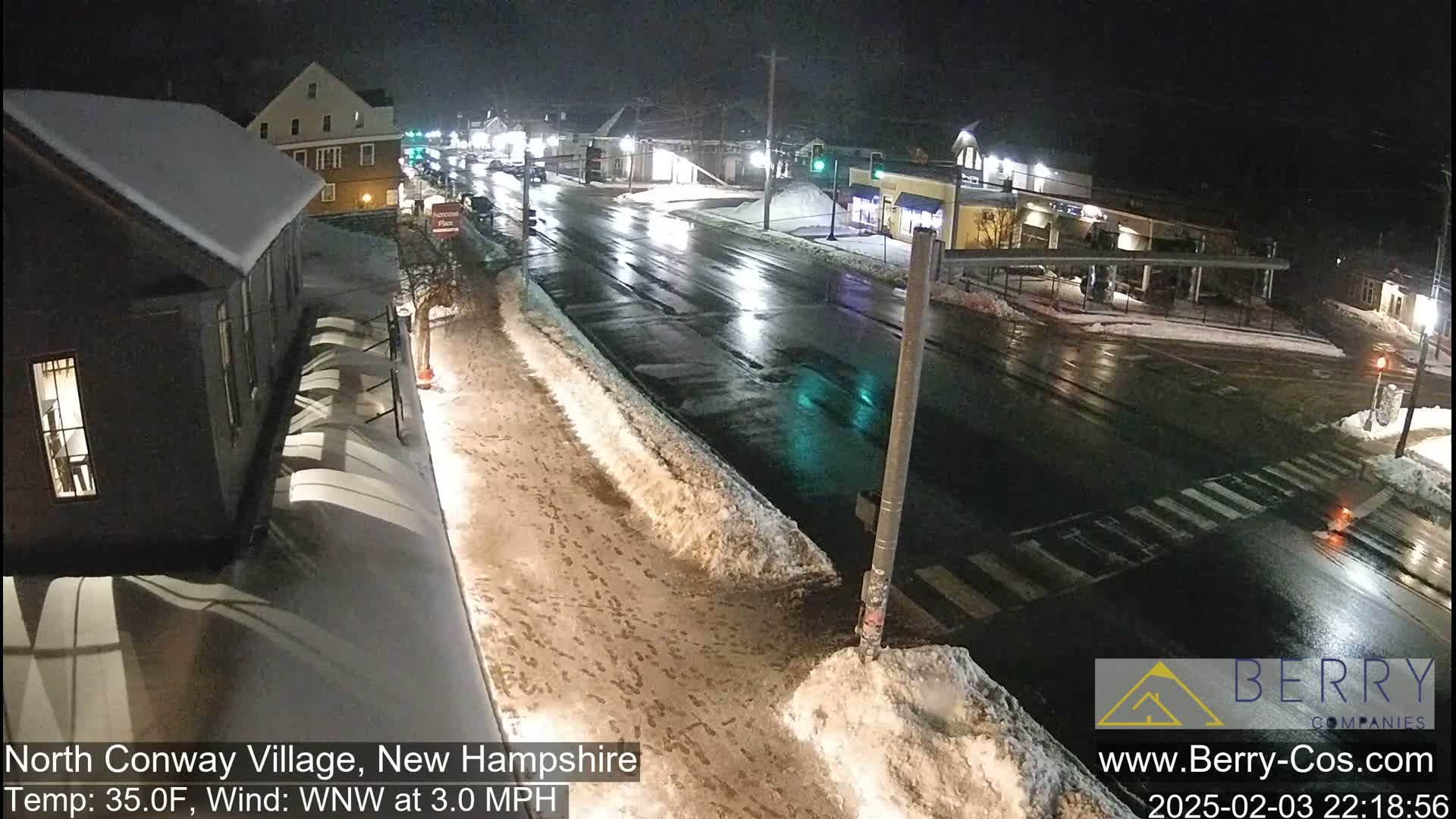 A nighttime street scene shows a wet road with snow banks on the sides, under partly cloudy skies.