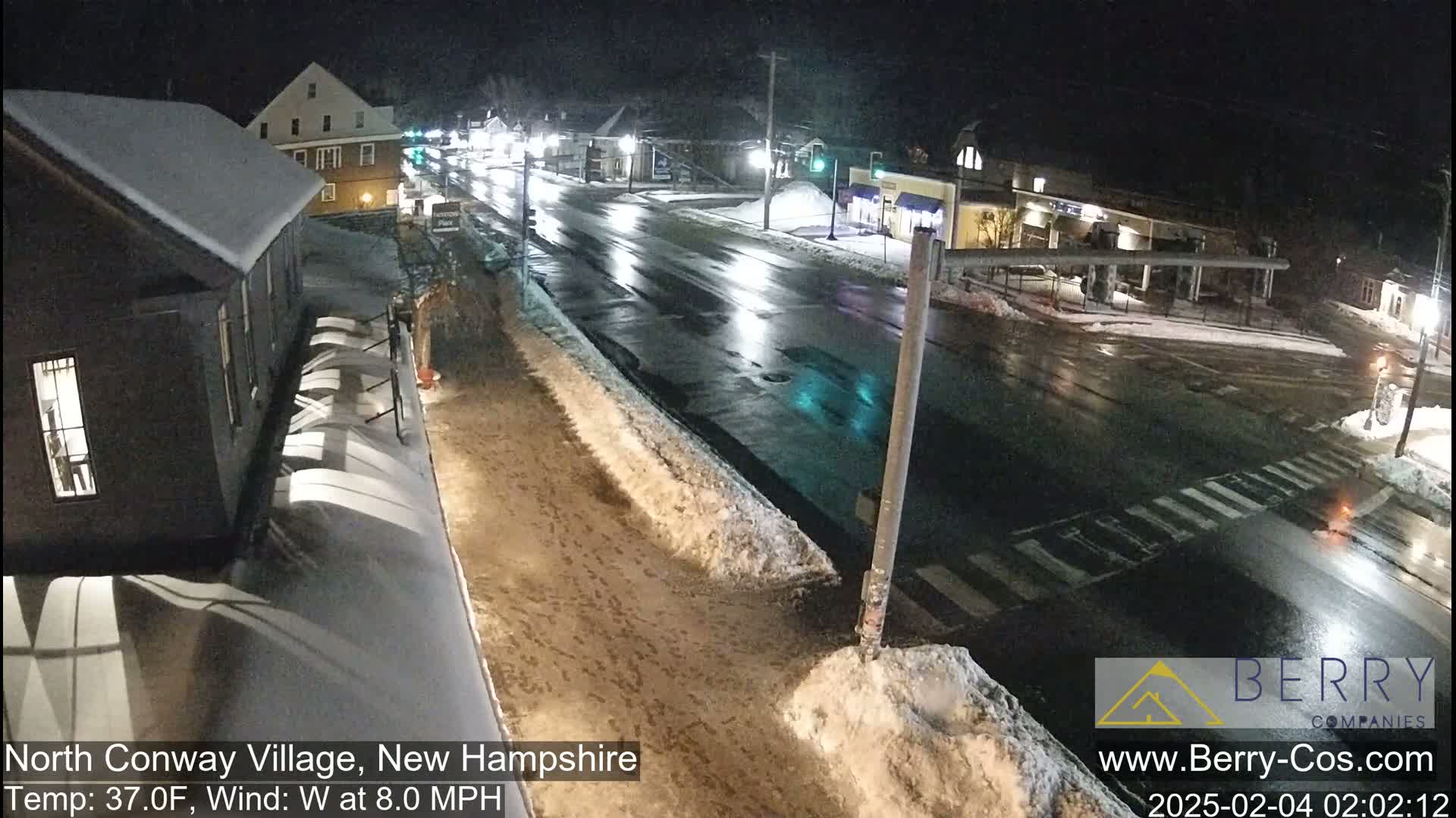 A nighttime view of a snow-covered street scene with wet roads and buildings under partly cloudy skies.