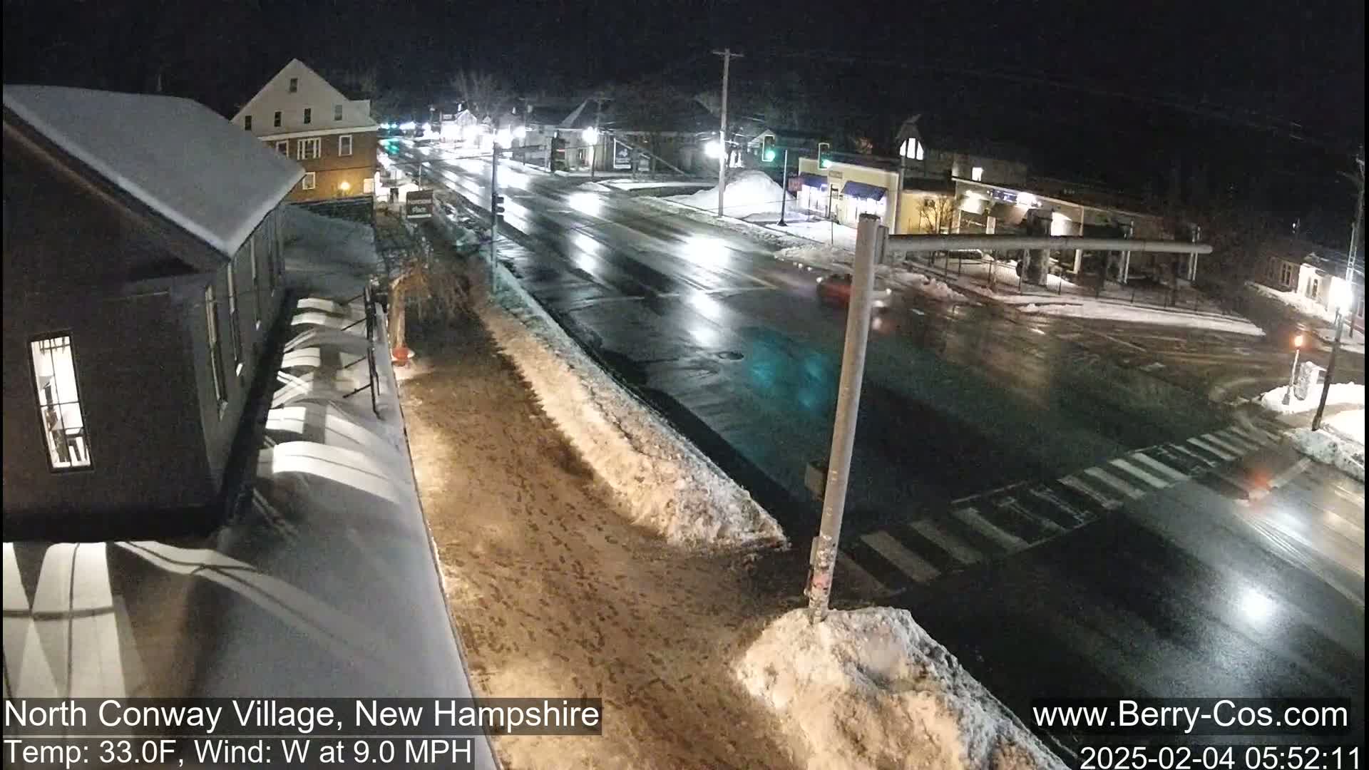 A nighttime view of a snow-covered street intersection with several buildings and a few cars, under partly cloudy skies.