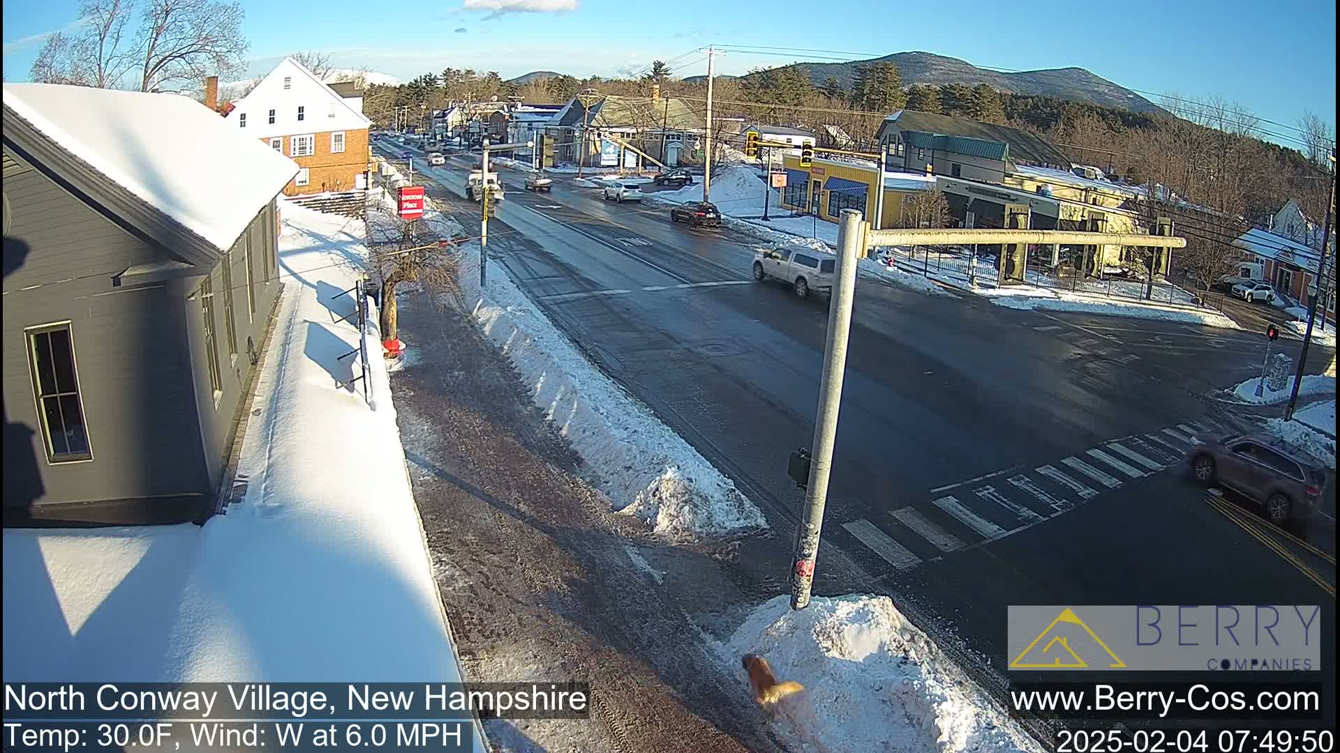 A snow-covered street scene shows a village intersection with several cars and buildings, under a sunny sky.