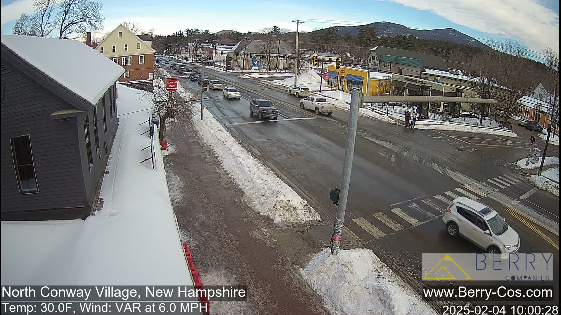 A snow-covered street scene shows several vehicles at an intersection in a small town, under a partly sunny sky.