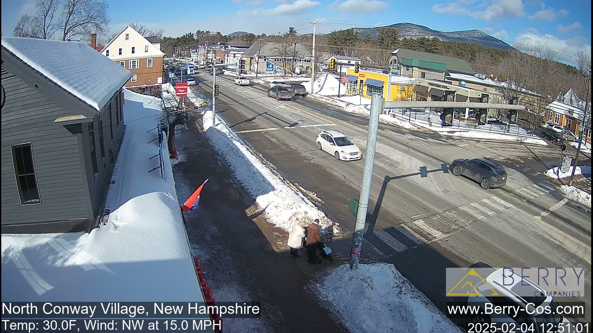 A snow-covered street scene shows several vehicles driving on a road that intersects with a crosswalk where pedestrians are walking, and buildings and mountains are visible in the background under a sunny sky.
