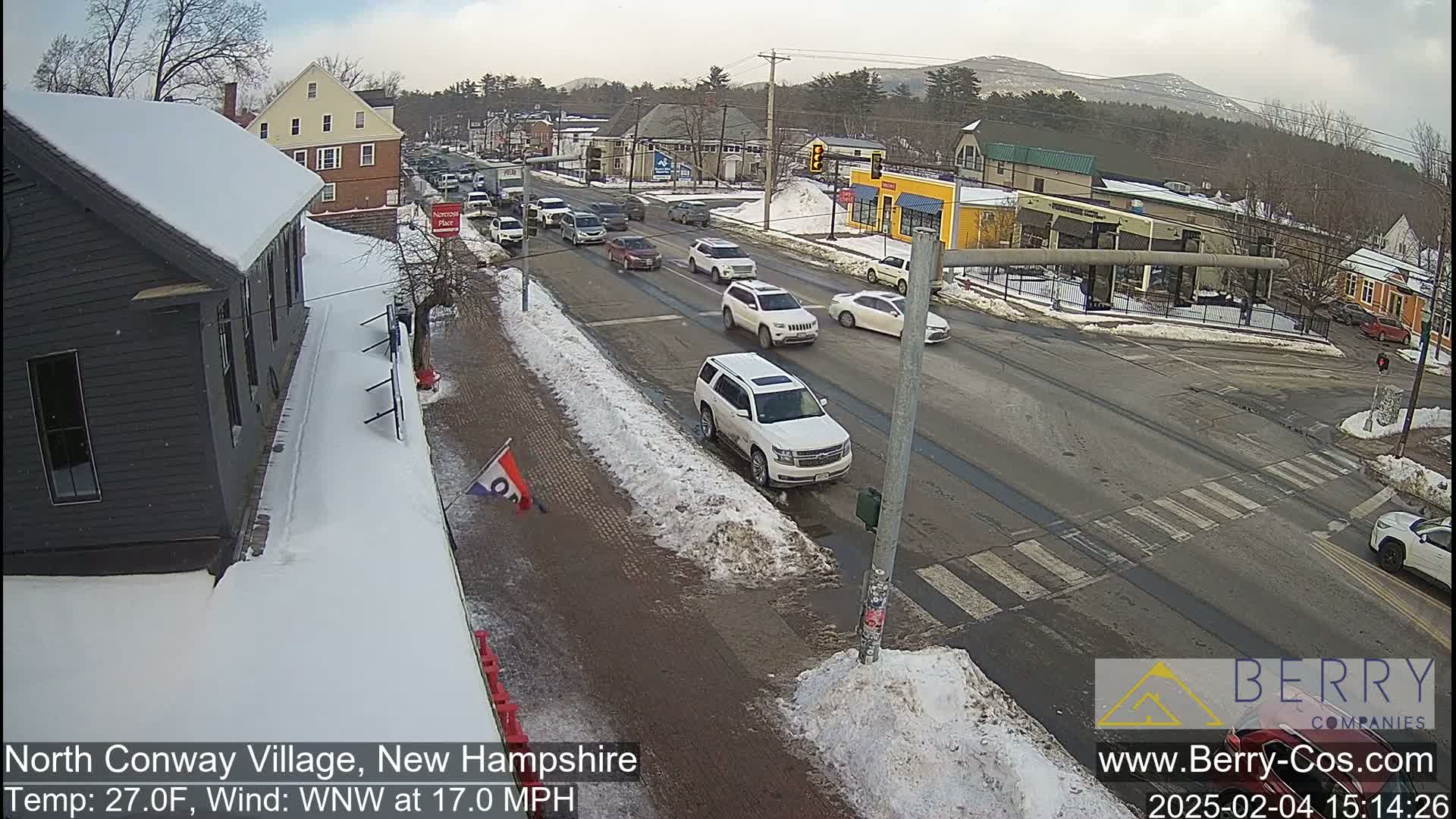 A snow-covered street scene shows several vehicles driving on a road that passes in front of small buildings, under a partly cloudy sky.