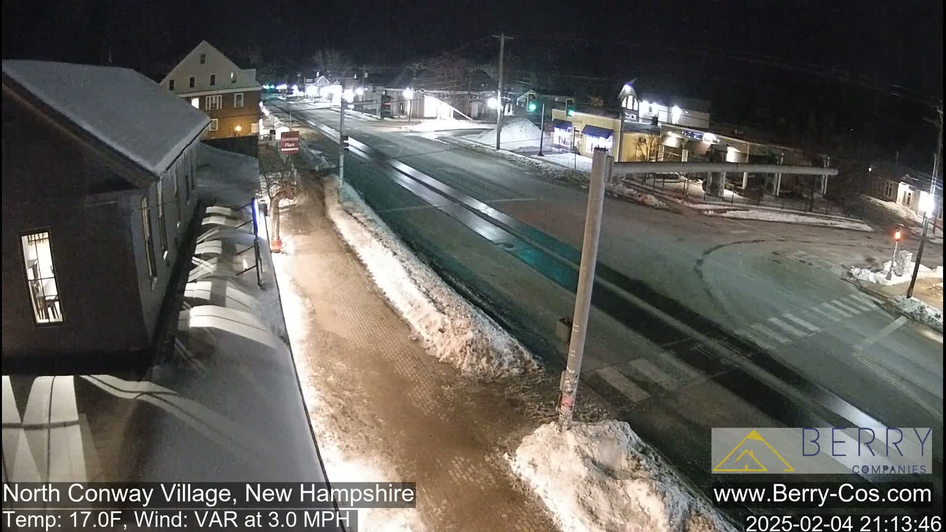 A nighttime view of a snow-covered street in a small town, with some buildings visible and a few cars on the road.