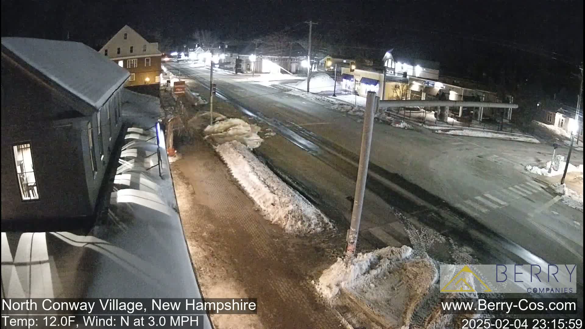 A snow-covered street scene at night shows buildings and a road intersection with partly melted snow and ice on the ground.
