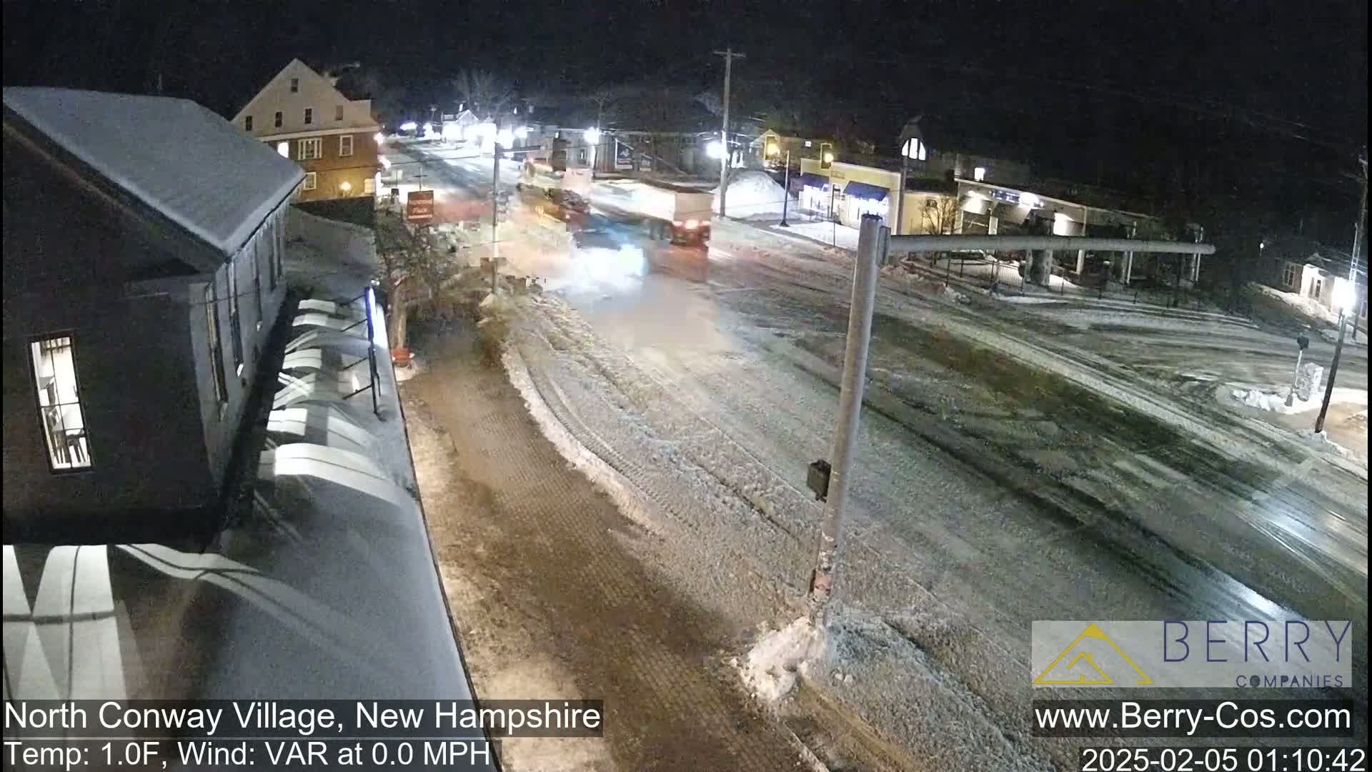 A snow-covered street at night with a few vehicles, lit by streetlights, with buildings lining the road.