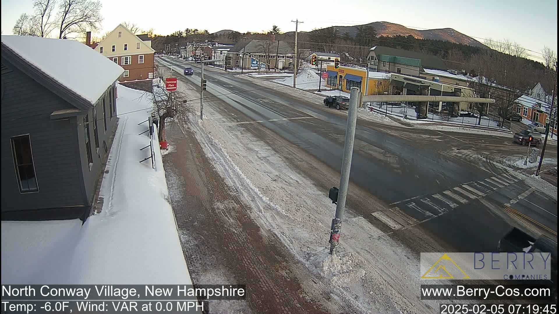 A snow-covered street scene shows a road intersection with several buildings, some cars, and a partly cloudy sky.