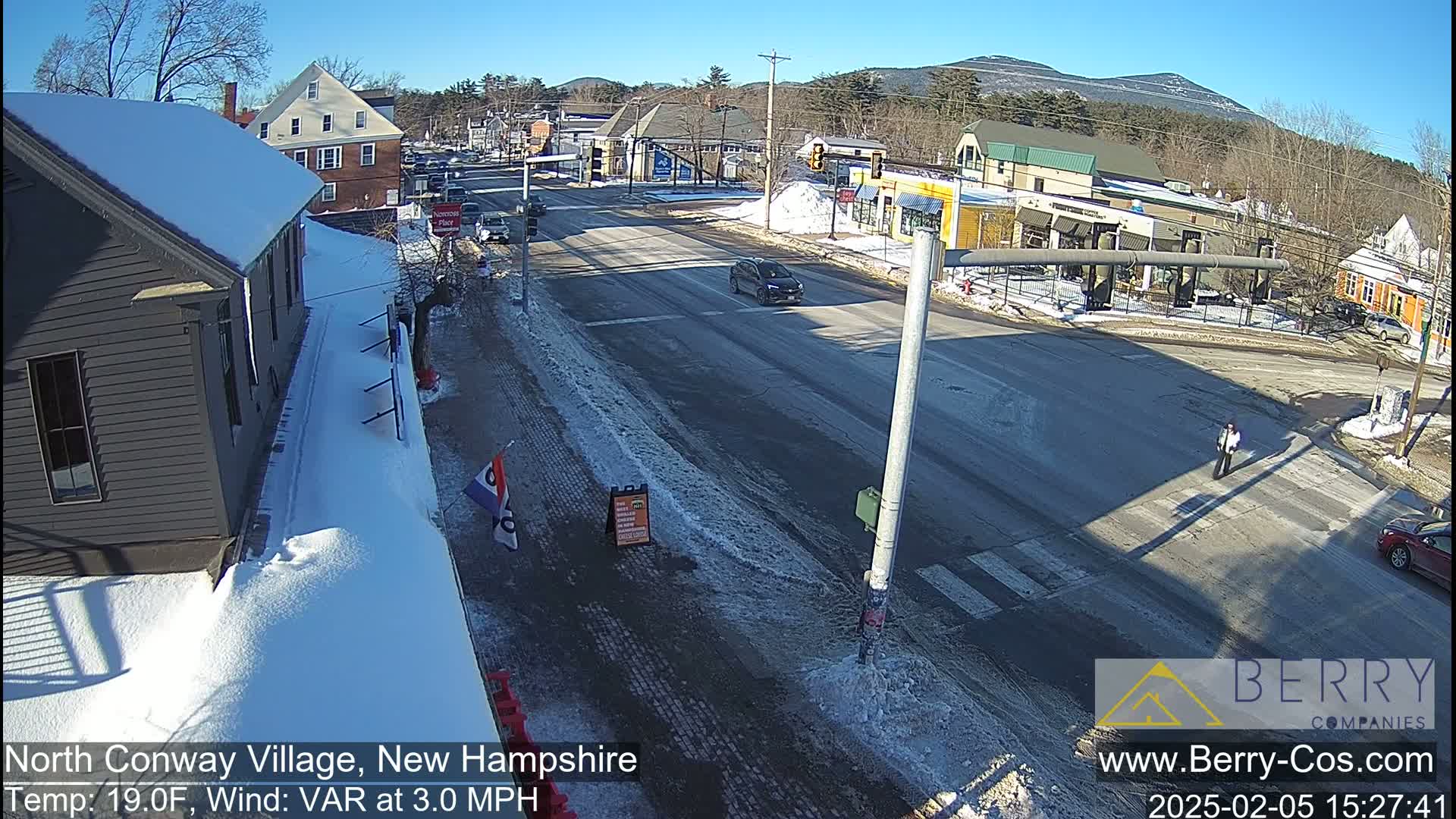 A snow-covered street scene shows a village intersection on a sunny day with a few cars and pedestrians.