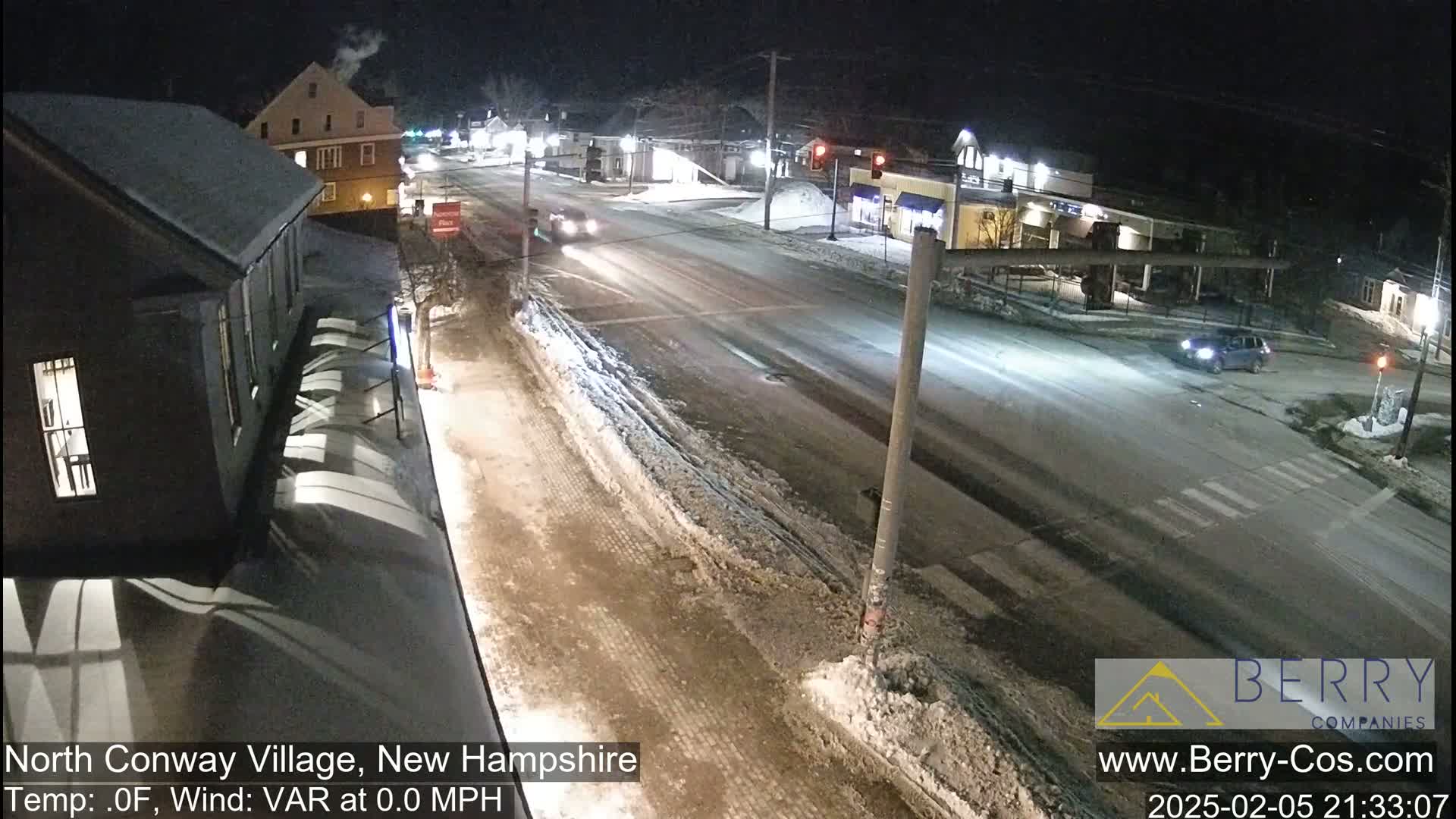 A snow-covered street scene at night, showing a two-lane road with a few cars, buildings on either side, and minimal traffic.