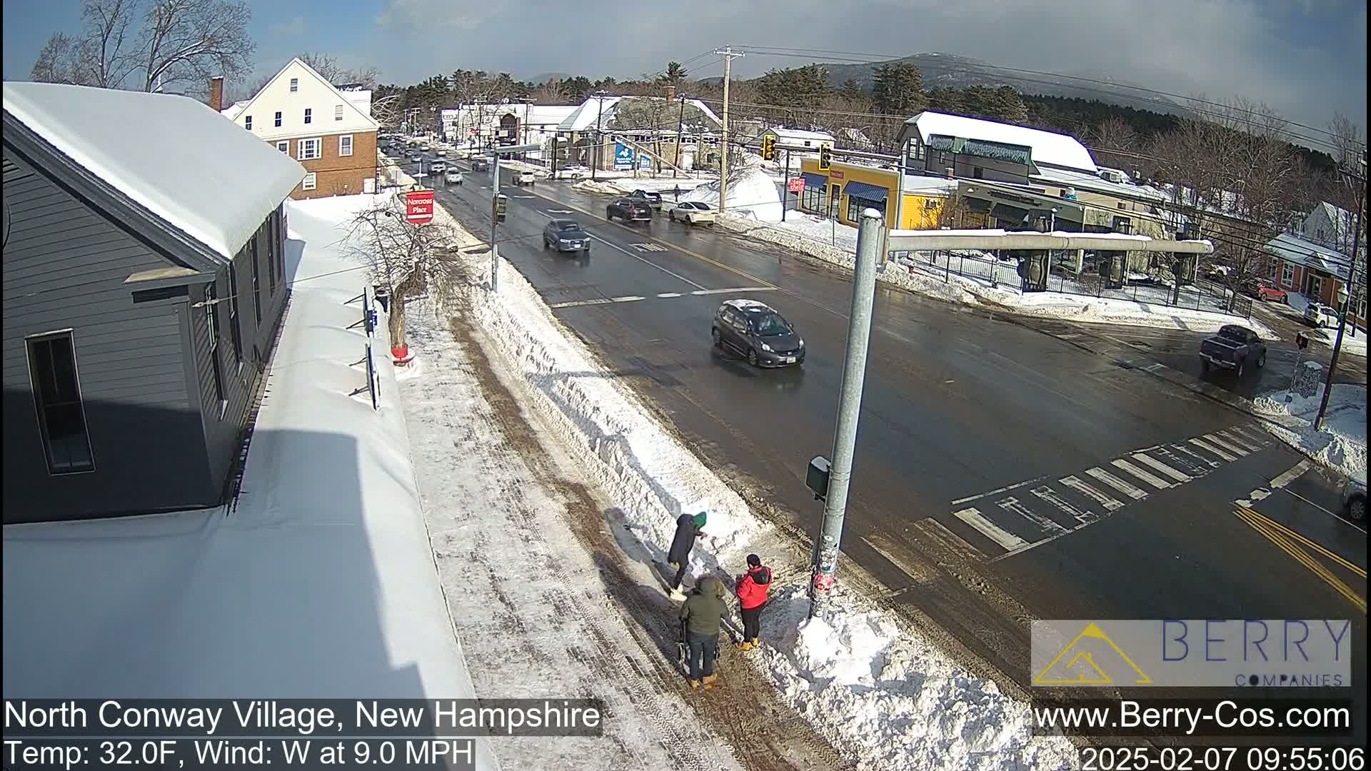 A sunny winter day shows a snow-covered street scene with several cars driving on a wet road, and people shoveling snow near a building.
