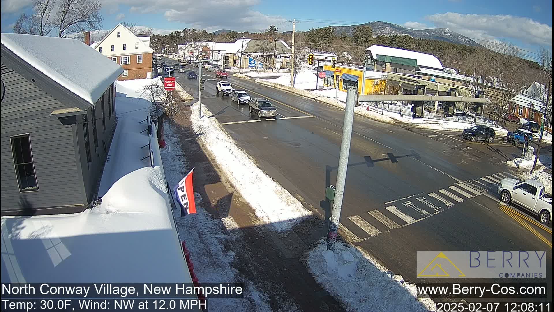 A partly sunny winter day shows a snow-covered street intersection in a small town with several cars driving by, and buildings on either side of the road.